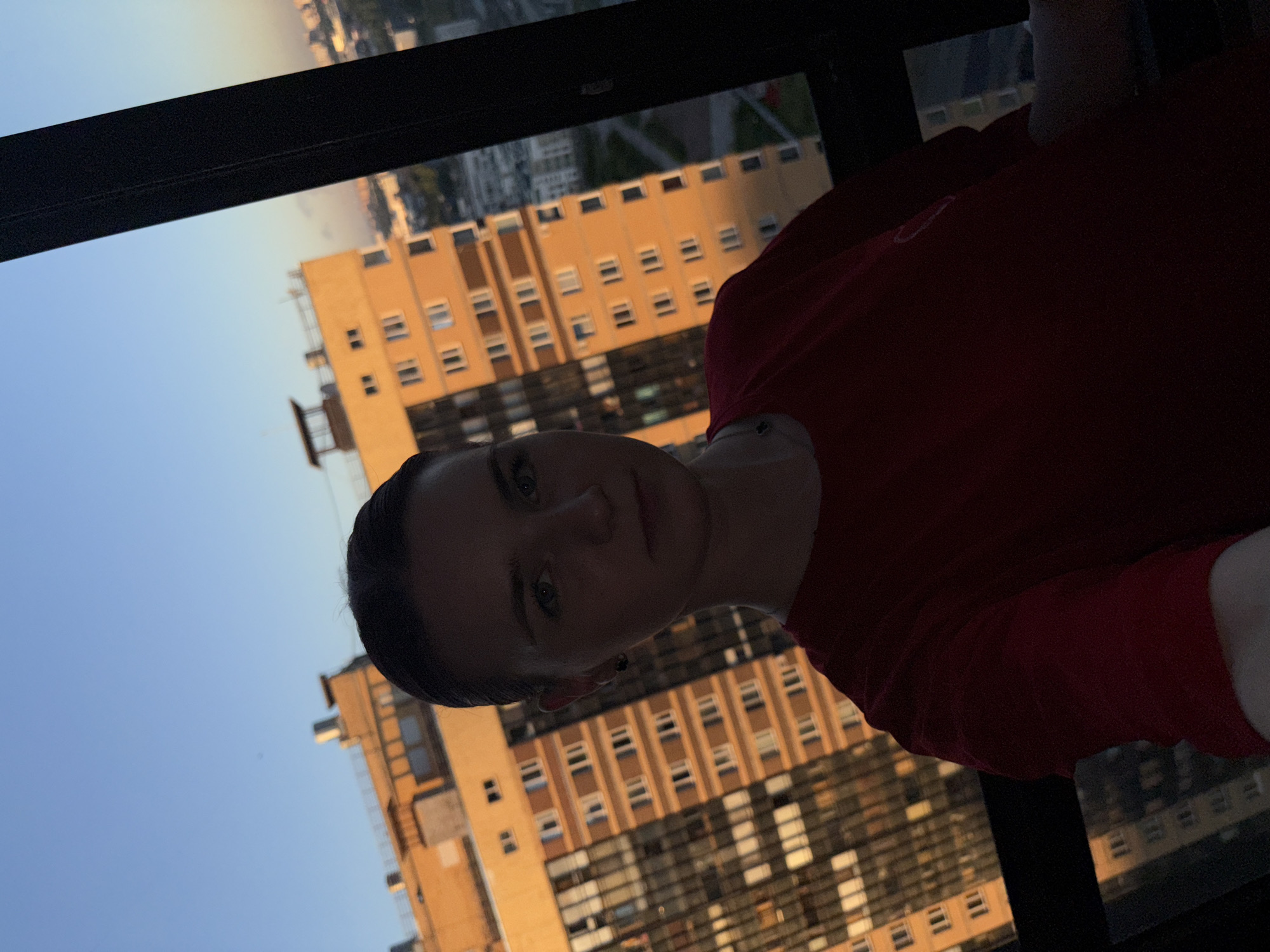 A young woman with dark hair pulled back from her face is taking a selfie. She is wearing a red t-shirt and a necklace. She is standing in front of a window, looking out at a city skyline at sunset. The buildings are tall and modern, and the sky is a clear blue with a hint of orange from the setting sun. The scene appears to be from a high-rise apartment or office building.