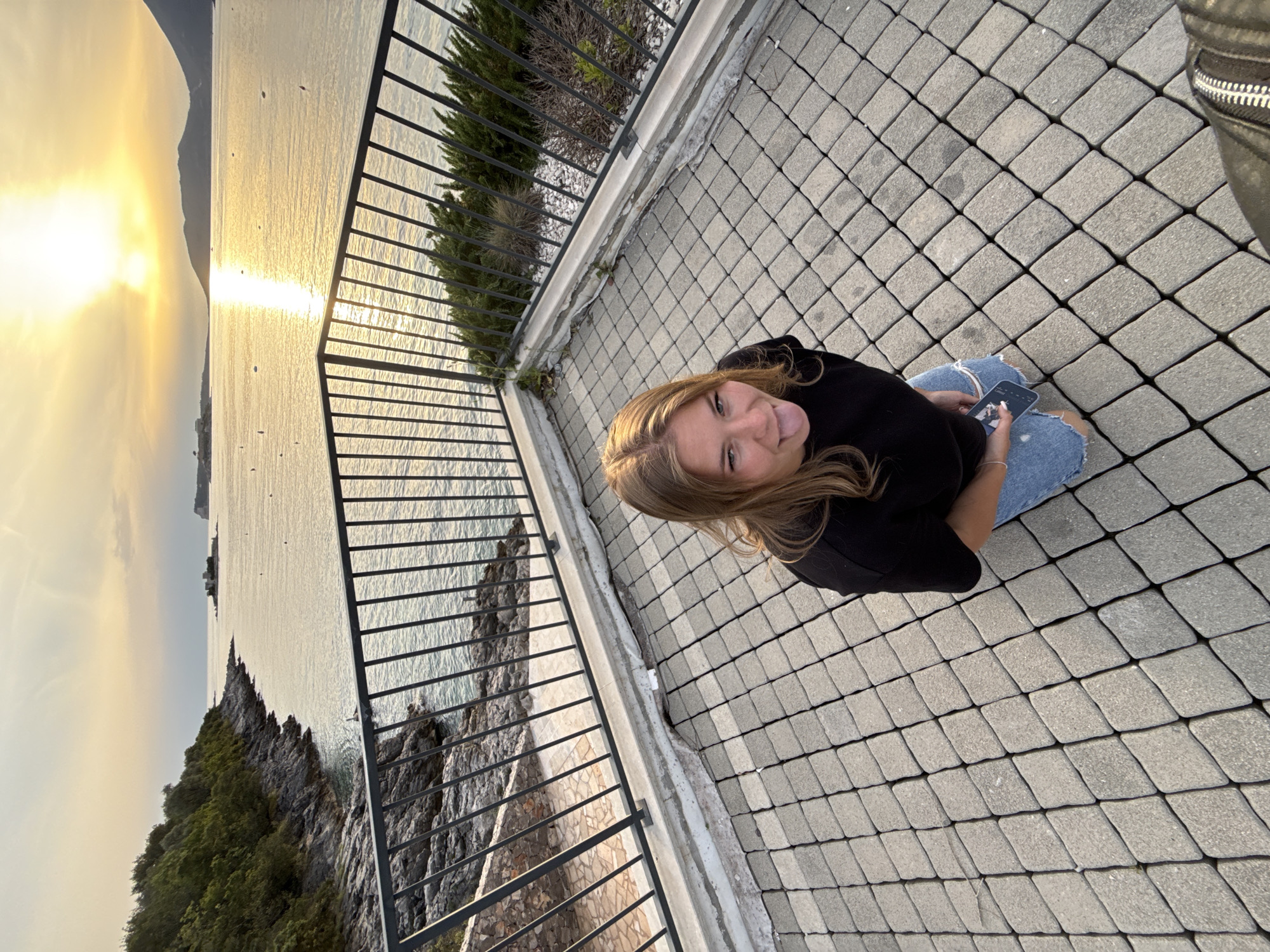 A woman with blonde hair and a black top is sitting on a paved surface. She is holding a cell phone. In the background, there is a body of water, possibly the Adriatic Sea, with land visible on the far side, and a bright sunset. A black metal fence runs along the left side of the frame. The setting appears to be a paved terrace or walkway overlooking the water. The location is likely in or near Luštica, Montenegro, given the landscape and coastal setting.