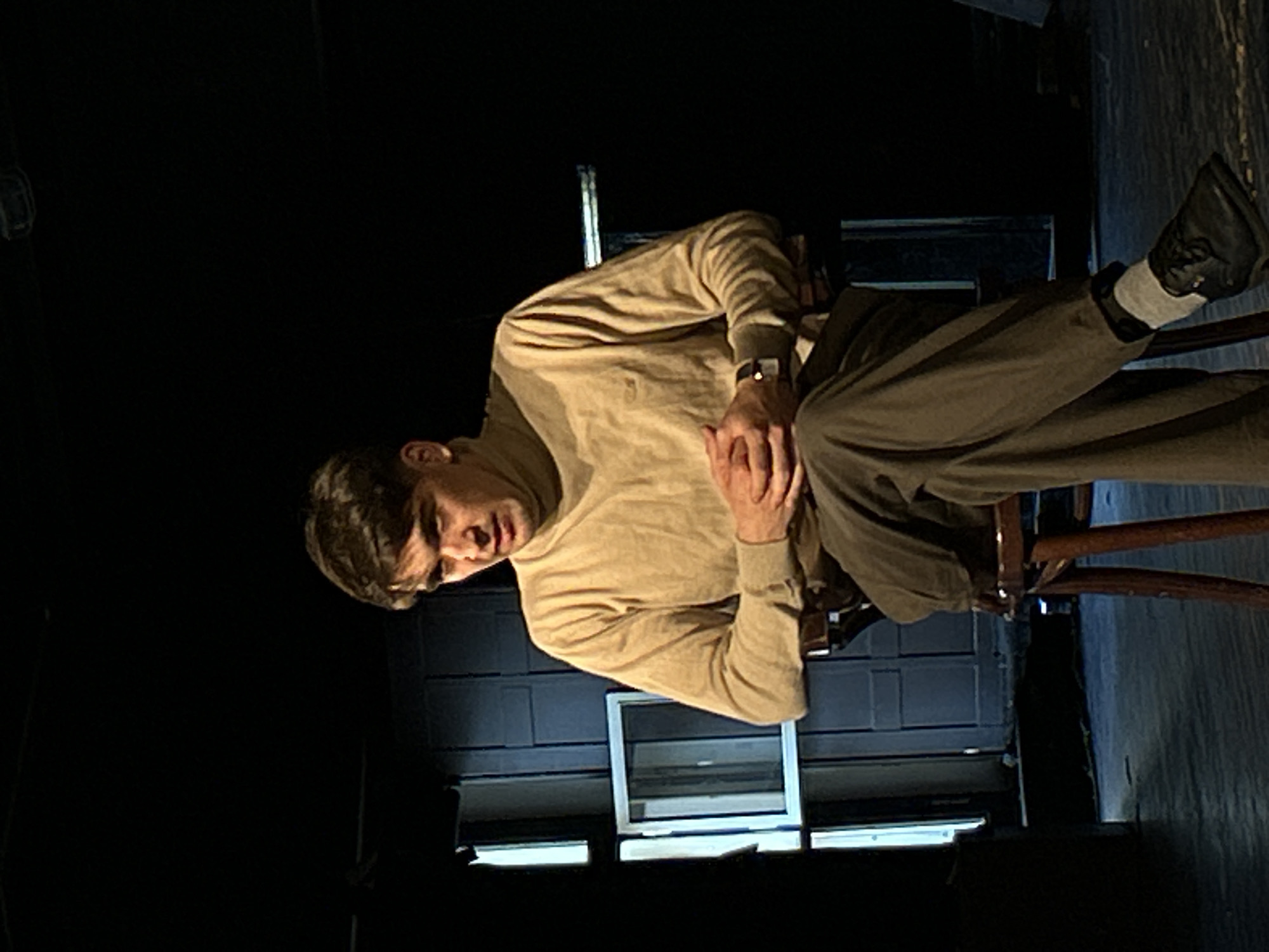 A man is seated on a wooden chair in a dimly lit, dark indoor setting. He is oriented as if sitting upright, although the image itself is rotated. The man has short, dark hair and is wearing a light beige or tan long-sleeved turtleneck sweater, dark trousers, and dark shoes with light-colored socks. A watch with a dark strap is visible on his left wrist.
He is leaning forward slightly, with his hands clasped together in front of him, resting on his lap. His gaze is directed forward or slightly downward, and his expression appears intense, focused, or possibly serious. The man is well-lit, suggesting a spotlight or deliberate artificial lighting, which contrasts sharply with the very dark background.
Behind him, faint rectangular shapes are visible, which could be windows or part of a set design, hinting at a theatrical stage or a sparsely furnished room. The floor beneath the chair appears to be dark wood. The overall mood conveyed by the lighting and the man's posture is dramatic or contemplative. No text is visible in the image.