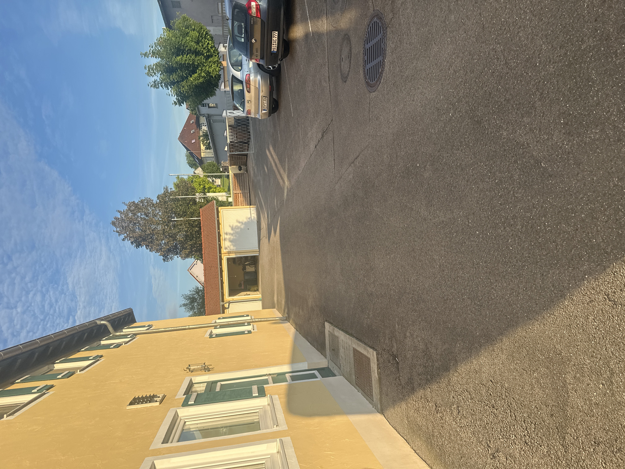 The image captures a sunlit scene in Mainburg, Germany, likely in the late afternoon or early evening. In the foreground, the yellow facade of a building is illuminated, casting a long shadow across the asphalt-paved area. The building features several windows with green shutters. Adjacent to the building, a rectangular patch of concrete with two manhole covers is visible.
Further into the scene, a low-slung building with a red-tiled roof houses a garage with two white doors, one of which is open. Inside the garage, various items are stored, including a bicycle. Beyond the garage, a metal fence with a diamond pattern encloses an area.
Several cars are parked in the paved area. A silver hatchback with the license plate "KEH C 306" is parked facing away from the camera. To its right, a dark-colored hatchback with the license plate "MAI AC 777" is also parked.
The background reveals a residential area with other buildings, trees, and a bright blue sky with scattered white clouds. The overall impression is one of a quiet, residential street bathed in the warm glow of the setting sun.
