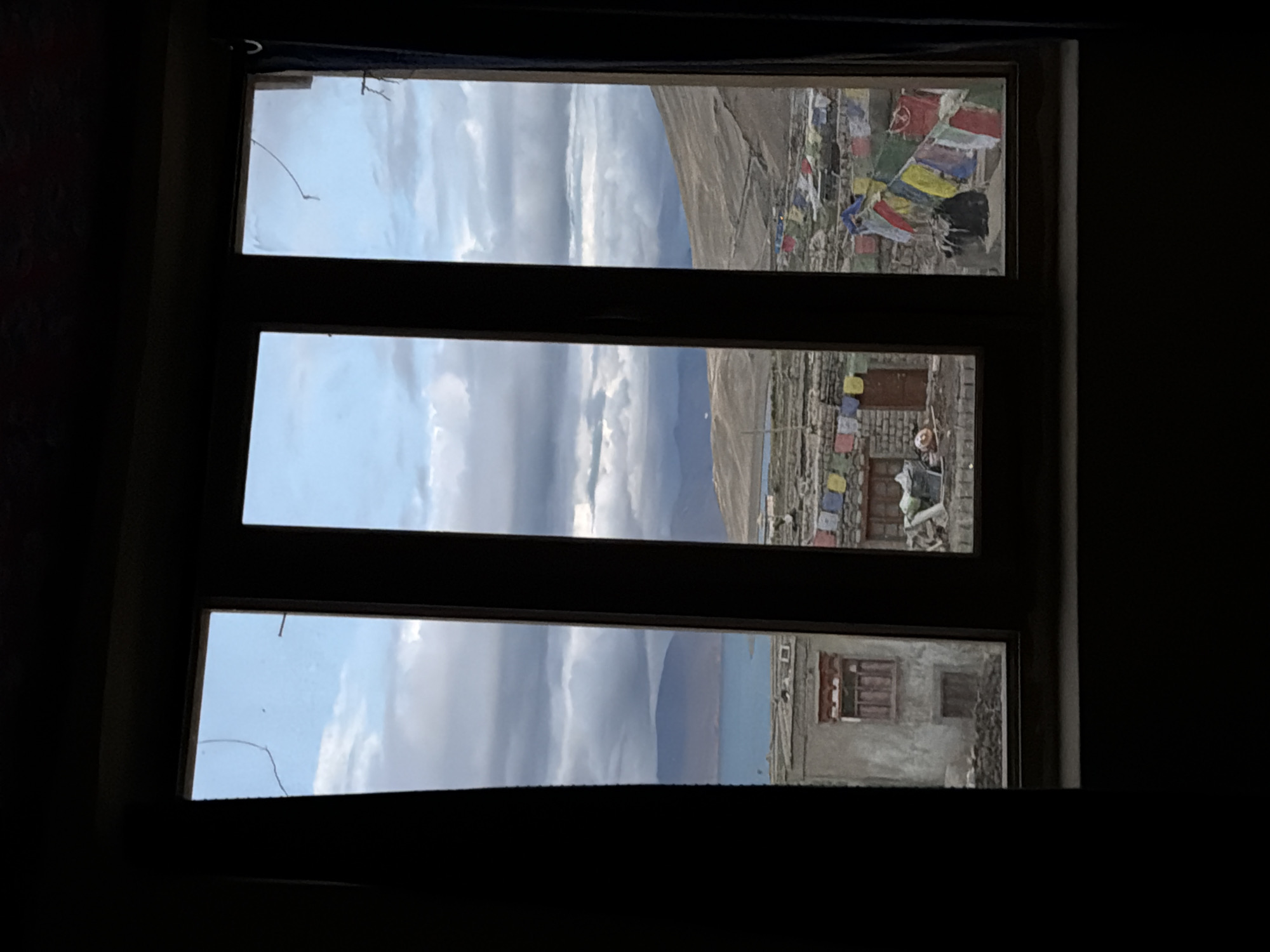 The image is a view through a three-pane window looking out onto the village of Karzok, India, situated on the shore of Tso Moriri lake. The sky is overcast with dramatic clouds. In the foreground, through the window, are buildings with stone and plastered walls. Prayer flags, in various colors including white, red, blue, yellow, and green, are strung between the buildings. A body of water, identified as Tso Moriri lake, is visible in the middle ground, with mountainous terrain surrounding it. The overall scene depicts a rustic, high-altitude environment.