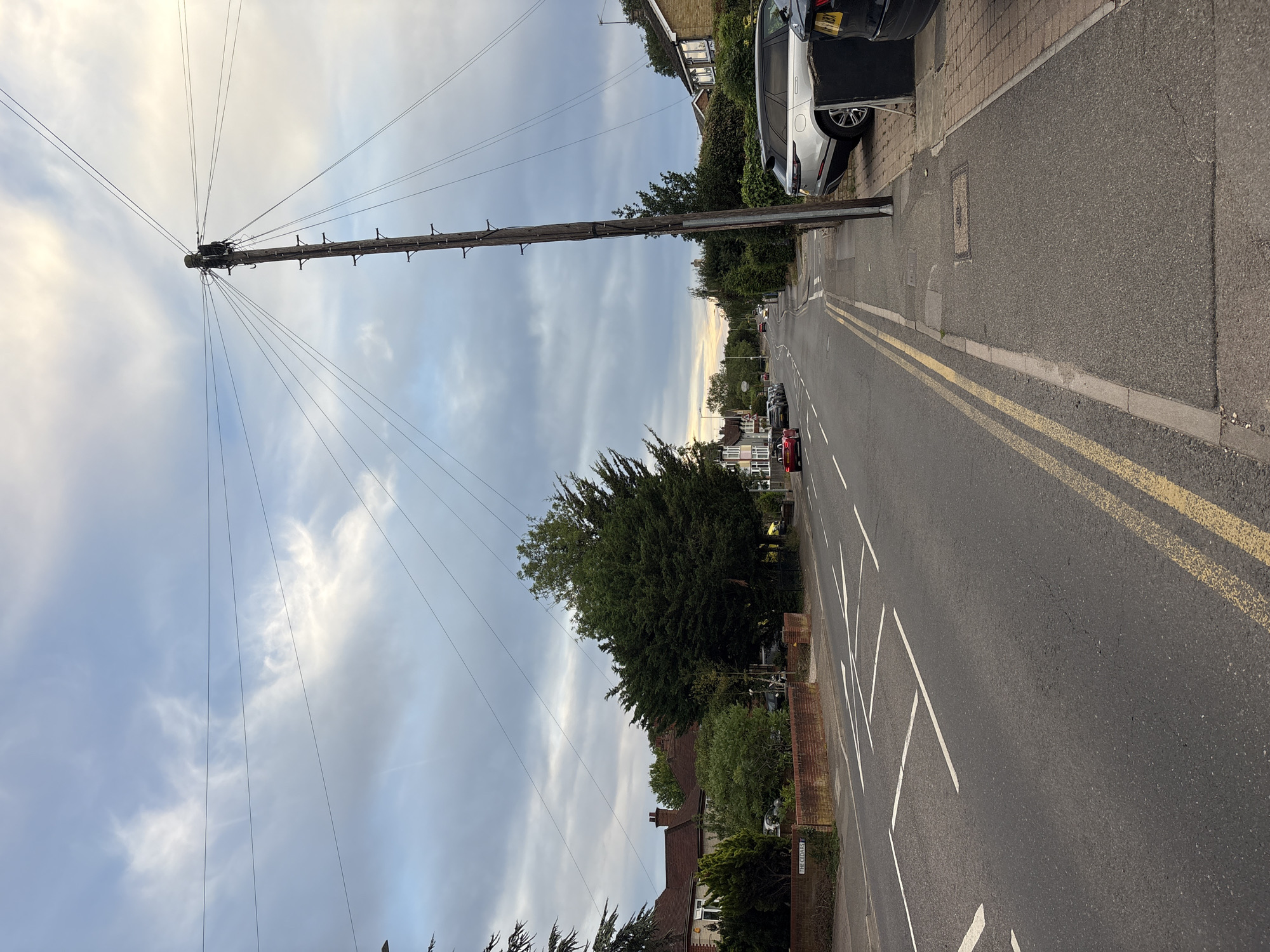 The image depicts a residential street scene in Chelmsford, United Kingdom, at dusk. A tall wooden utility pole with numerous wires extending from it dominates the right side of the frame. The street is asphalt, with double yellow lines marking the center and white road markings indicating a pedestrian crossing ahead. Several cars are parked along the side of the road, including a silver SUV. On the left side of the street, there are houses with brick walls and gardens. The sky is cloudy, with hints of sunset colors in the distance. A street sign labeled "THE CEDARS" is visible on the left.