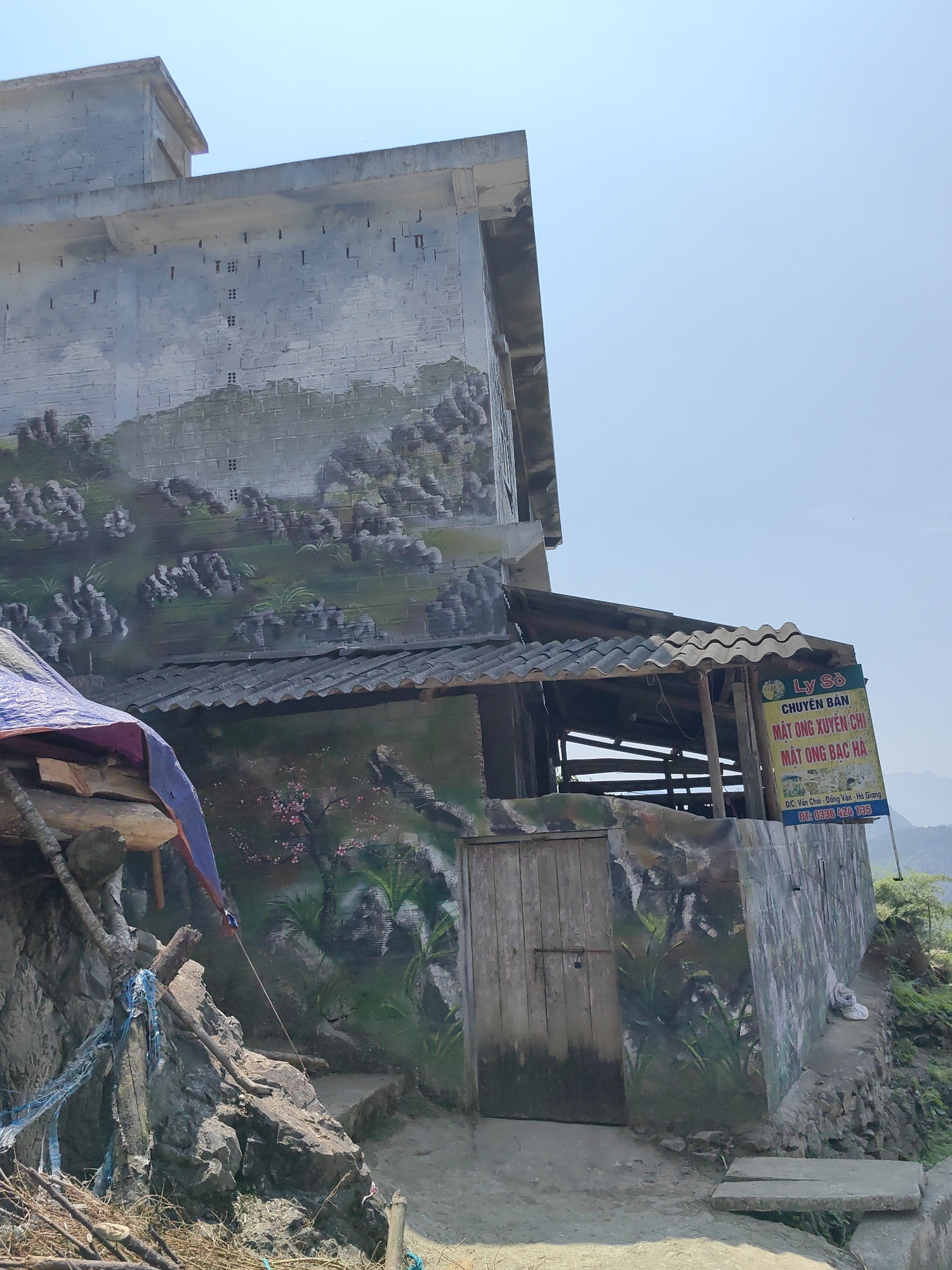 The image captures a building in Yên Minh Commune, Vietnam, adorned with landscape paintings on its exterior walls. The building is multi-level, with the upper section painted with a depiction of mountainous terrain. A lower section features a smaller wooden door, and the wall is painted with vegetation and flowers. An awning provides partial cover to the entrance area. A sign in Vietnamese, advertising honey products, is attached to the building's side. The foreground includes a rocky slope with some wooden posts, branches, and blue rope. The sky is visible in the background and appears bright and clear.