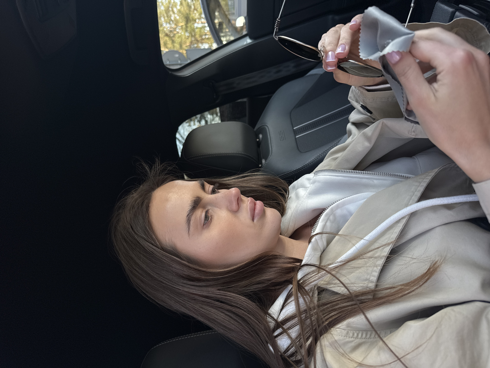 A woman is sitting in a car cleaning a pair of sunglasses with a cleaning cloth. She is wearing a white hoodie under a light brown trench coat. The car interior is black with gray seats. The location cannot be identified as Białobrzegi, Poland based on the image.
