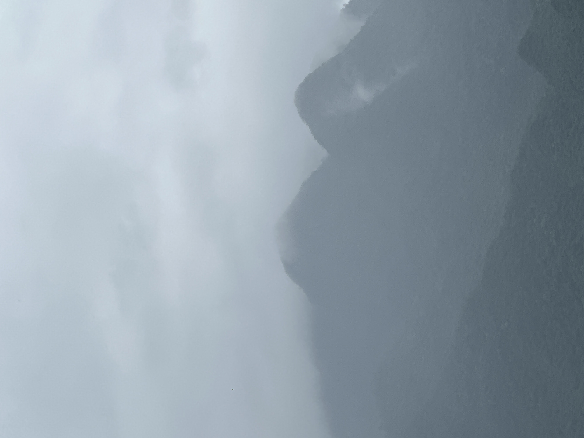 The image shows a misty mountain landscape. Several mountain peaks are visible, shrouded in fog and clouds. The foreground consists of lower, darker hills covered in dense vegetation. The sky is overcast and gray, with wispy clouds. There are no visible people or man-made structures. The scene is tranquil and appears to be a natural landscape. The location is Xóm Khán, Vietnam, which is known for its mountainous terrain and lush greenery.