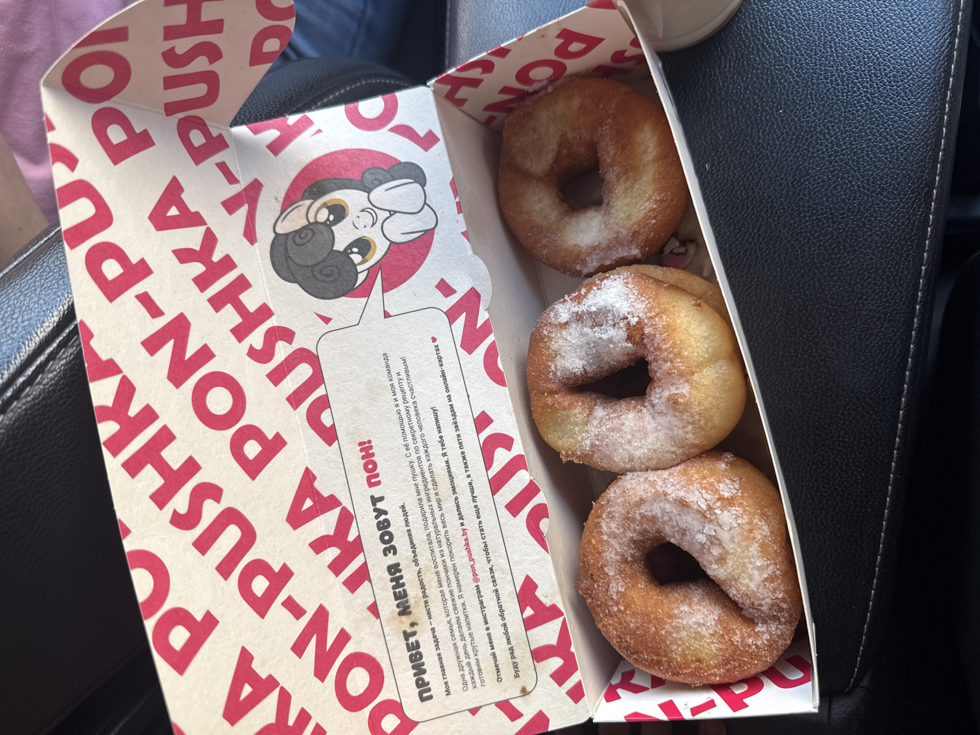 The image shows a cardboard box filled with three donuts. The box has red and white branding with the text "PON-PUSHK" and "PON-PUXKA PO" repeated. Inside the box, a cartoon pug character is depicted. The donuts are golden brown and sprinkled with sugar. One donut is in the top right, another in the bottom right, and a third is partially visible at the top of the box. The setting appears to be inside a car, with a black leather car seat visible to the right of the box. The lighting suggests it is daytime. A message in Russian is printed on the side of the box, starting with "ПРИВЕТ, МЕНЯ ЗОВУТ ПОН!" which translates to "HELLO, MY NAME IS PON!". The message is about the donuts bringing joy and being made with natural ingredients. The location context indicates Mahilyow, Belarus.