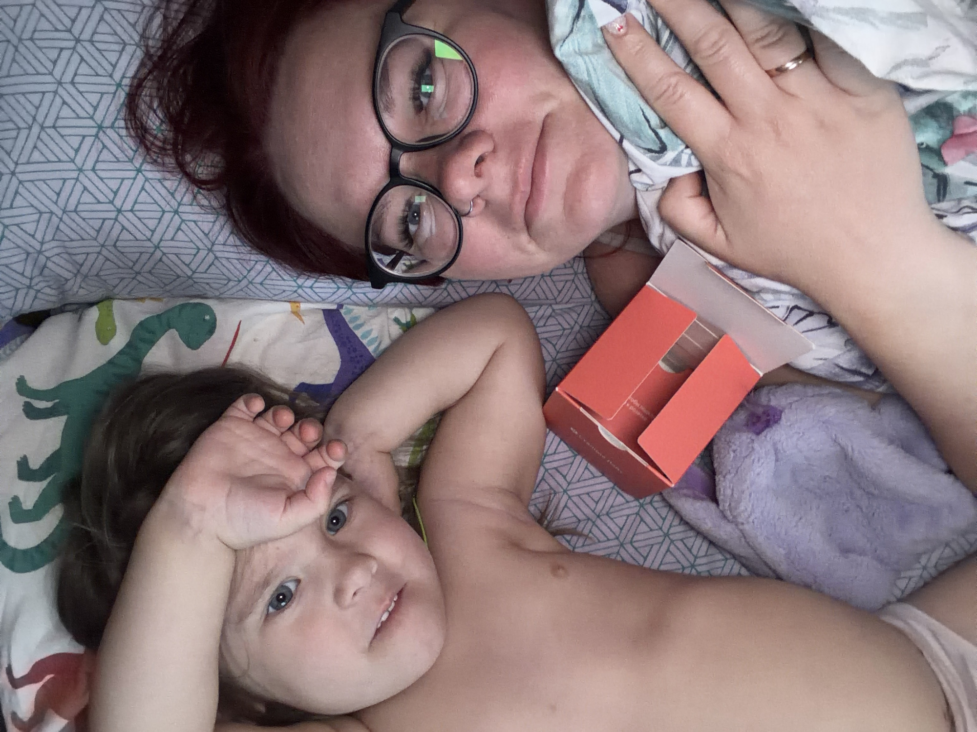 An adult female and a young child are lying side-by-side in a bed in Chaikovsky, Russia. The adult, with reddish-brown hair, is on the right, wearing black-rimmed glasses and a nose ring. Her right hand, wearing a gold band on the ring finger, holds a light green and blue patterned sheet. The child, on the left, has dark brown hair and blue eyes, is shirtless, and has a light-colored garment covering her lower torso. Her right arm is bent, with the hand resting on her forehead. Both individuals are looking directly forward. Between them, an open, empty red cardboard box is positioned on a pillow with a light blue geometric pattern. A purple plush object is partially visible beneath the red box and the child's arm. The child is resting on bedding featuring green dinosaur patterns.