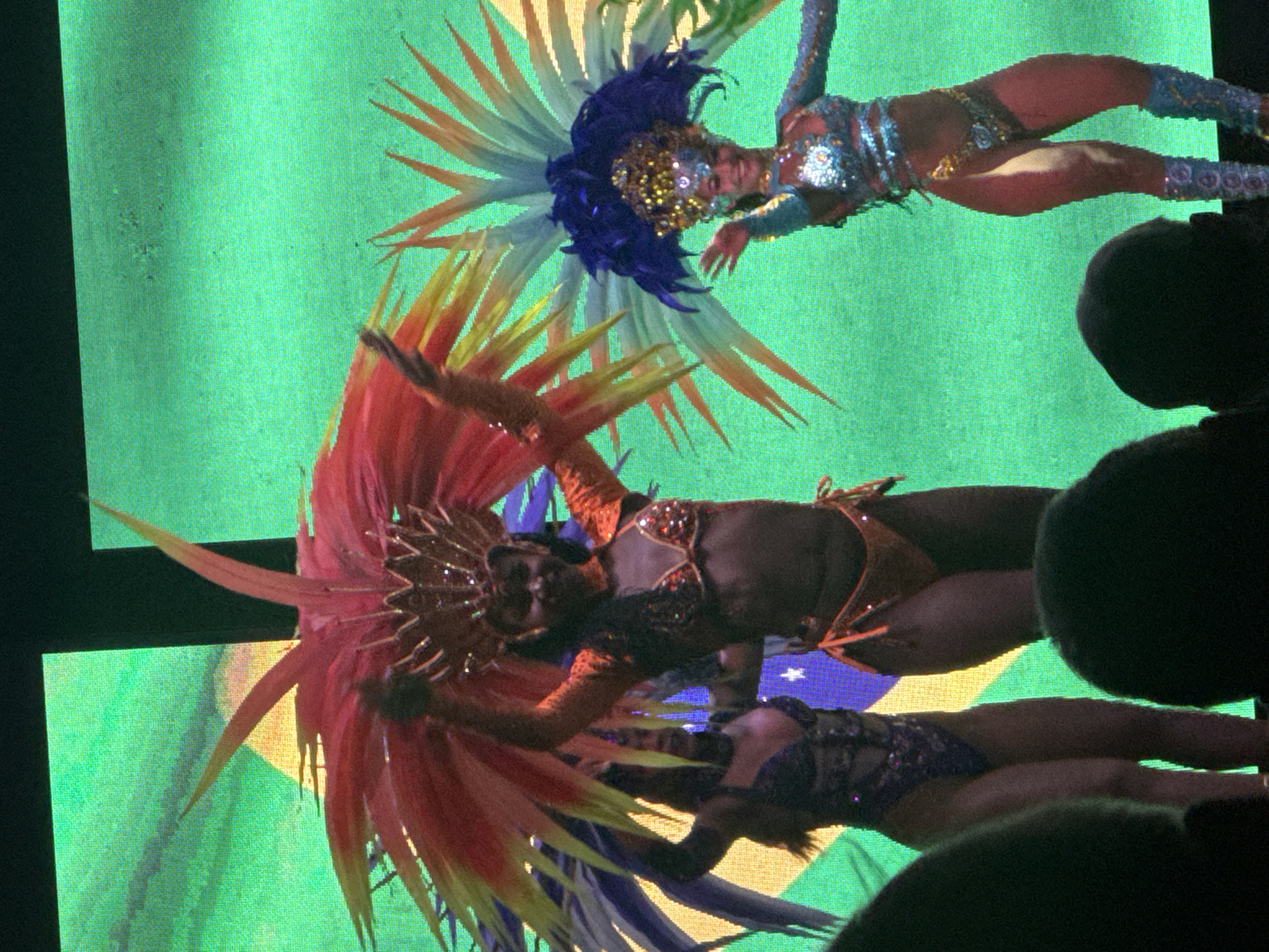 Three women in elaborate, colorful costumes are performing on a stage with a large green screen backdrop that displays a stylized Brazilian flag. The woman in the center has a large, fiery red and yellow headdress. The woman on the left wears a dark, jeweled costume and a headdress with blue, yellow, and green plumes. The woman on the right has a bright blue and yellow headdress and a shimmering blue and silver costume adorned with sequins. They appear to be part of a parade or a performance, possibly in Manavgat, Turkey, given the context provided. The image is framed from the perspective of an audience member, with the dark silhouettes of heads in the foreground.