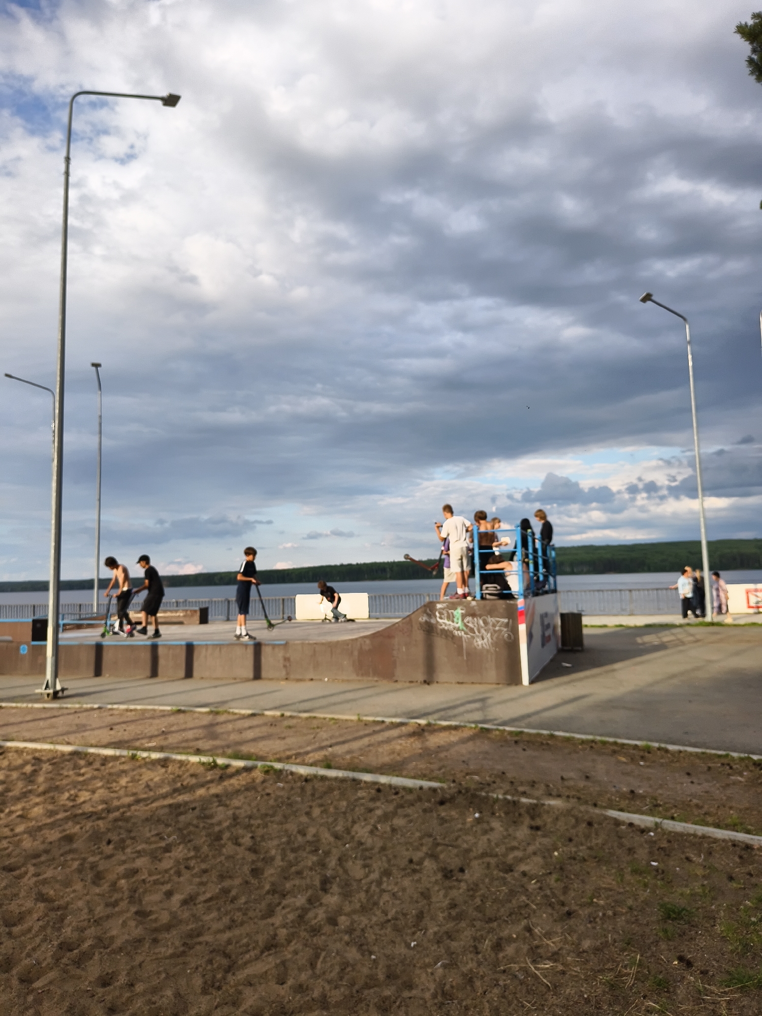 This image captures an outdoor scene at a skatepark in Chaikovsky, Russia. The skatepark features ramps and platforms, with several individuals actively engaged in activities such as skateboarding and scootering. One person is on a scooter, another appears to be preparing to ride down a ramp, and a group of people are standing on a raised platform. In the background, a body of water, likely a river or lake, is visible, with a treeline on the opposite shore. The sky is filled with dramatic, cloudy formations. Streetlights are positioned throughout the area. The foreground shows a sandy area, characteristic of a park or recreational space.