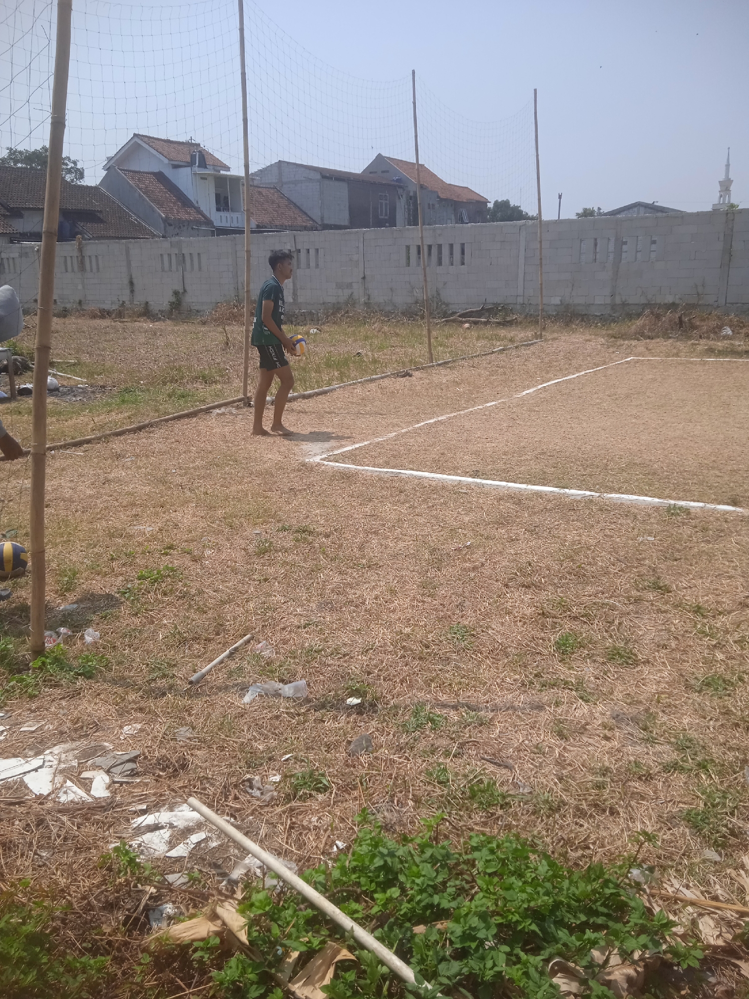 An outdoor, unkempt volleyball court is pictured in Klodran, Indonesia. The court is defined by white lines on a brown, grassy surface. A volleyball net is suspended between two bamboo poles. A young man in a green t-shirt and black shorts is standing on the court, holding a volleyball. He is barefoot. In the background, there are houses and a concrete wall. The scene appears to be under a bright, clear sky. There is debris scattered on the ground around the court.