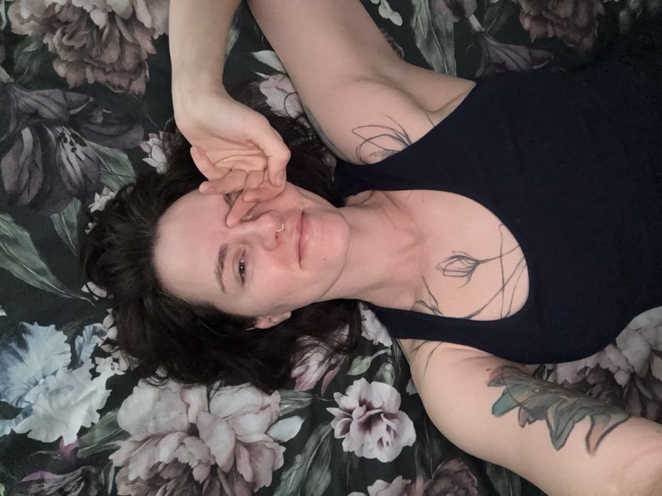 An adult female subject is lying supine on a bed adorned with a dark, floral-patterned bedspread featuring large, stylized light-colored flowers. The subject wears a dark sleeveless garment. Dark, medium-length hair frames the head. A silver ring is visible piercing the left nostril. The subject's right hand is positioned with fingers resting near the right temple. Visible tattoos include linear, plant-like motifs on the subject's right chest and right arm, and a multi-colored floral or organic design on the left upper arm. The subject's eyes are partially open, and lips form a slight smile. The scene is observed from an overhead perspective indoors, located in Boreti, Montenegro.