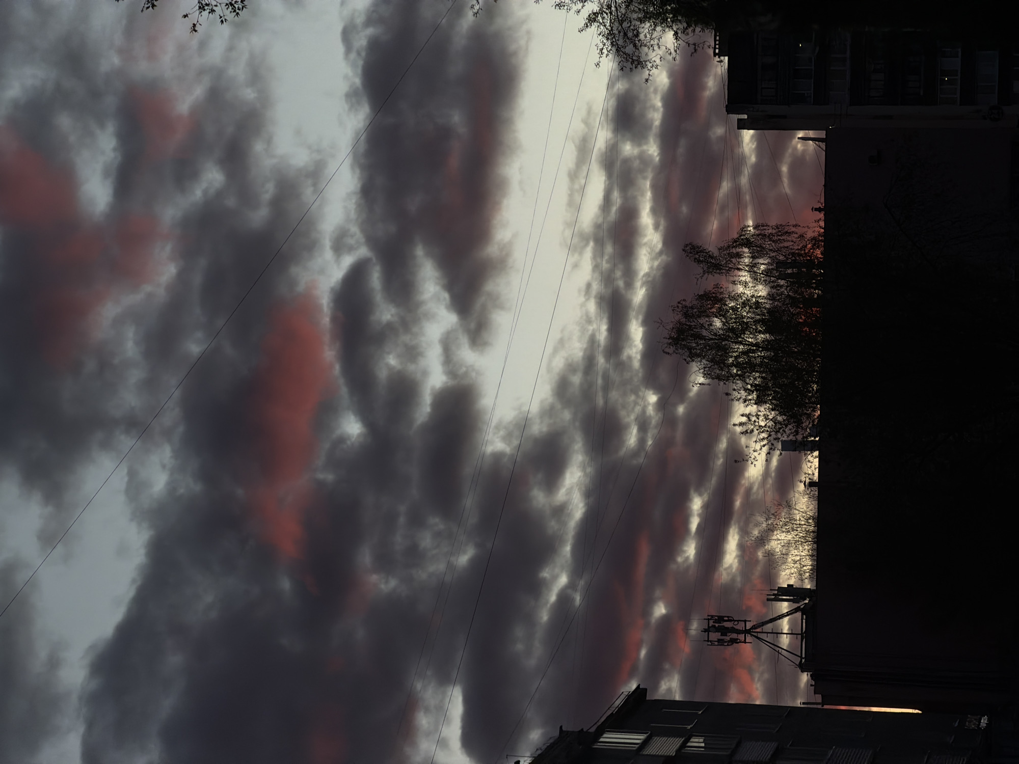 The image depicts a twilight or sunset scene in a cityscape, likely in Perm, Russia, judging by the architectural style and overcast sky. The foreground features dark, heavy clouds with hints of red or orange coloration, possibly from the setting sun. Thin electrical wires stretch across the sky, intersecting the view of the clouds. Adjacent to the cloudy sky is a dark silhouette of a building, with bare tree branches visible against the sky. Power lines or utility poles and wires are attached to the building. The lower part of the image shows the tops of other buildings, with windows visible. The overall impression is a contrast between the natural beauty of the sky and the stark lines of urban infrastructure. The sky is positioned on the left.
