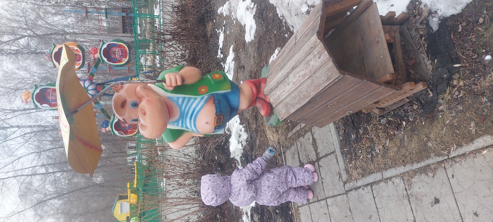 The image depicts an outdoor scene in what appears to be a park or playground setting, potentially located in Novosibirsk, Russia. There is a child wearing a purple snowsuit lying on a concrete surface, near a wooden structure resembling a small shed or playhouse. Next to the child, there's a large, cartoonish statue of a figure with a pig-like face, dressed in blue overalls and a green vest. In the background, a partially visible children's carousel is present. Patches of snow are visible on the ground, indicating a cold or winter season. Bare trees and dry vegetation are also noticeable.