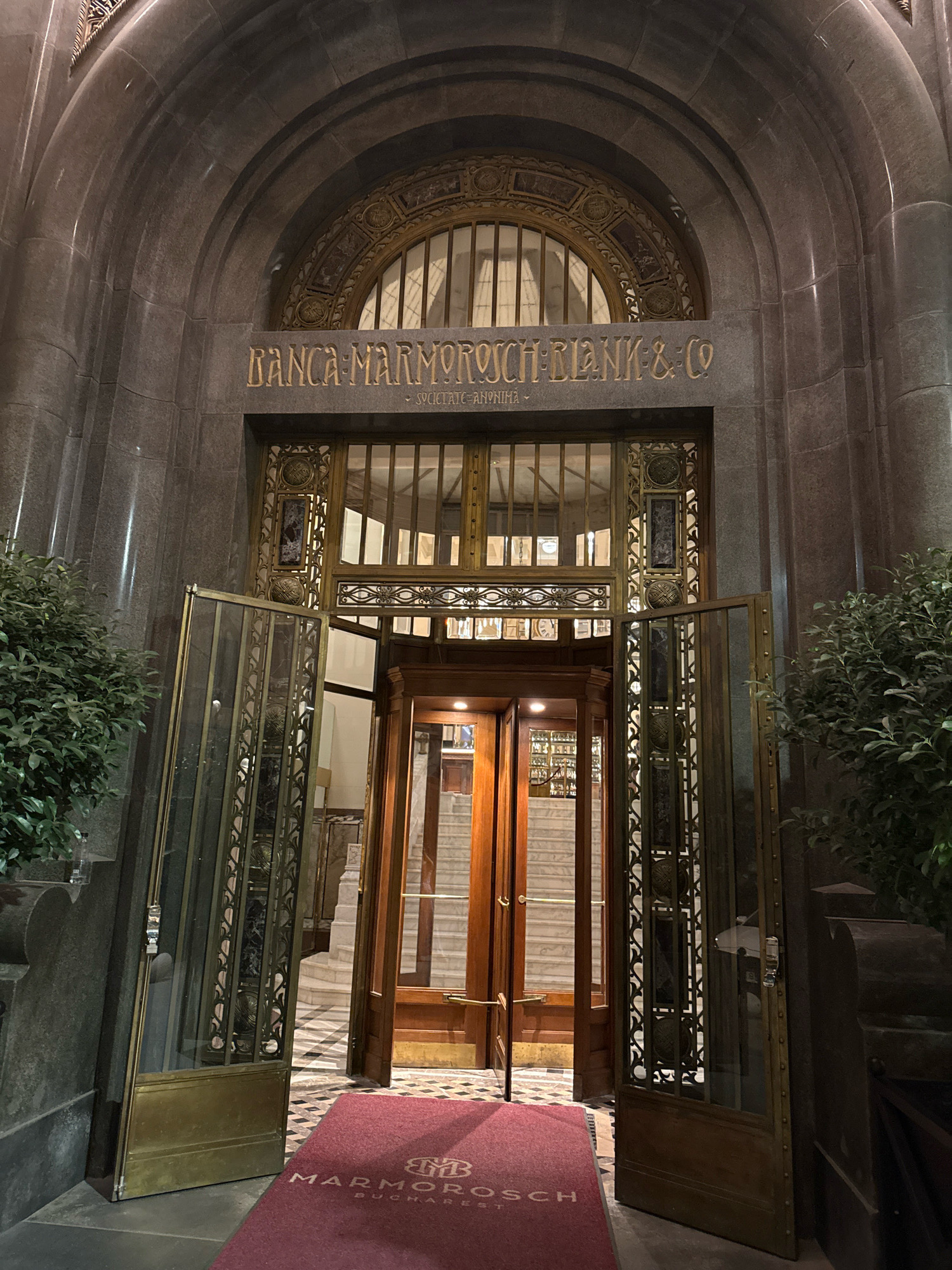 The image shows the entrance of the Marmorosch-Blank Bank building in Bucharest, Romania. The building's facade is made of dark stone, with a large arched entranceway. Above the entrance, the words "BANCA MARMOROSCH BLANK & CO" are inscribed in golden lettering, along with "SOCIETATE ANONYMA". The entrance itself features ornate brass and glass doors, with one door propped open. Inside, a grand staircase can be seen, with ornate railings and marble steps. A red carpet with the Marmorosch-Blank logo is placed on the ground in front of the entrance. Green bushes flank the entrance on either side. The lighting suggests it is nighttime or dusk, with artificial lights illuminating the entrance. There are no people visible in the image.