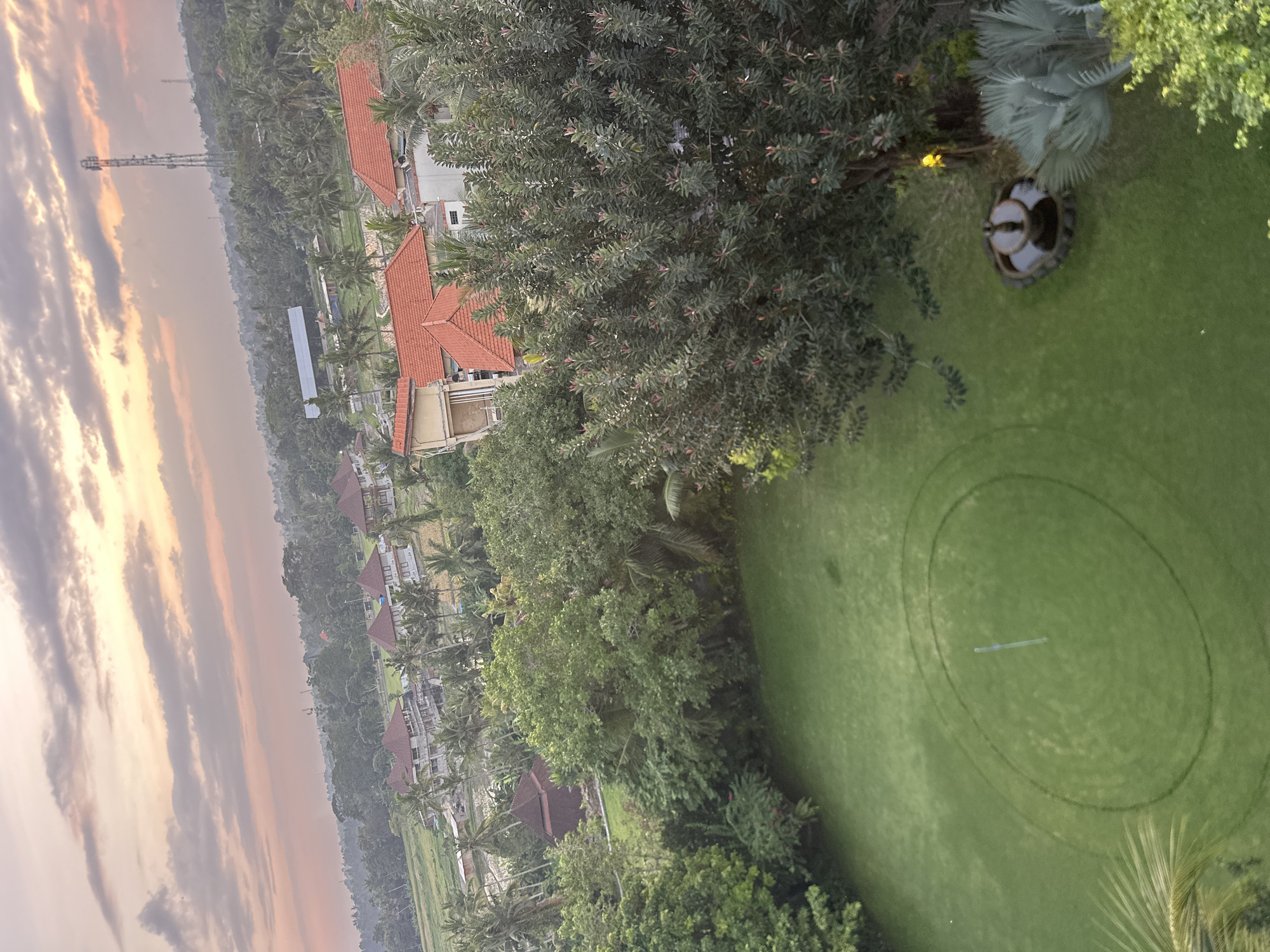 The image depicts a scenic view of a tropical landscape at dusk, likely in Ubud District, Indonesia. The sky is filled with dramatic, streaky clouds in shades of orange, pink, and gray, indicating either sunrise or sunset. In the foreground, a neatly manicured green lawn is visible, with two concentric circles mown into it, suggesting a practice area for a sport like golf or croquet. A light-colored, elongated object lies across the center of the circles, possibly a marker or a piece of equipment.
Beyond the lawn, a line of modern villas with red tile roofs is nestled amongst lush green trees and palm trees. Some of the villas appear to be multi-story structures. In the distance, a tall telecommunications tower stands against the horizon, surrounded by more dense vegetation. The overall impression is one of a peaceful, tropical resort or residential area, bathed in the soft, diffused light of twilight. The scene evokes a sense of tranquility and natural beauty, characteristic of a serene evening in a tropical paradise. No visible text is present in the image.