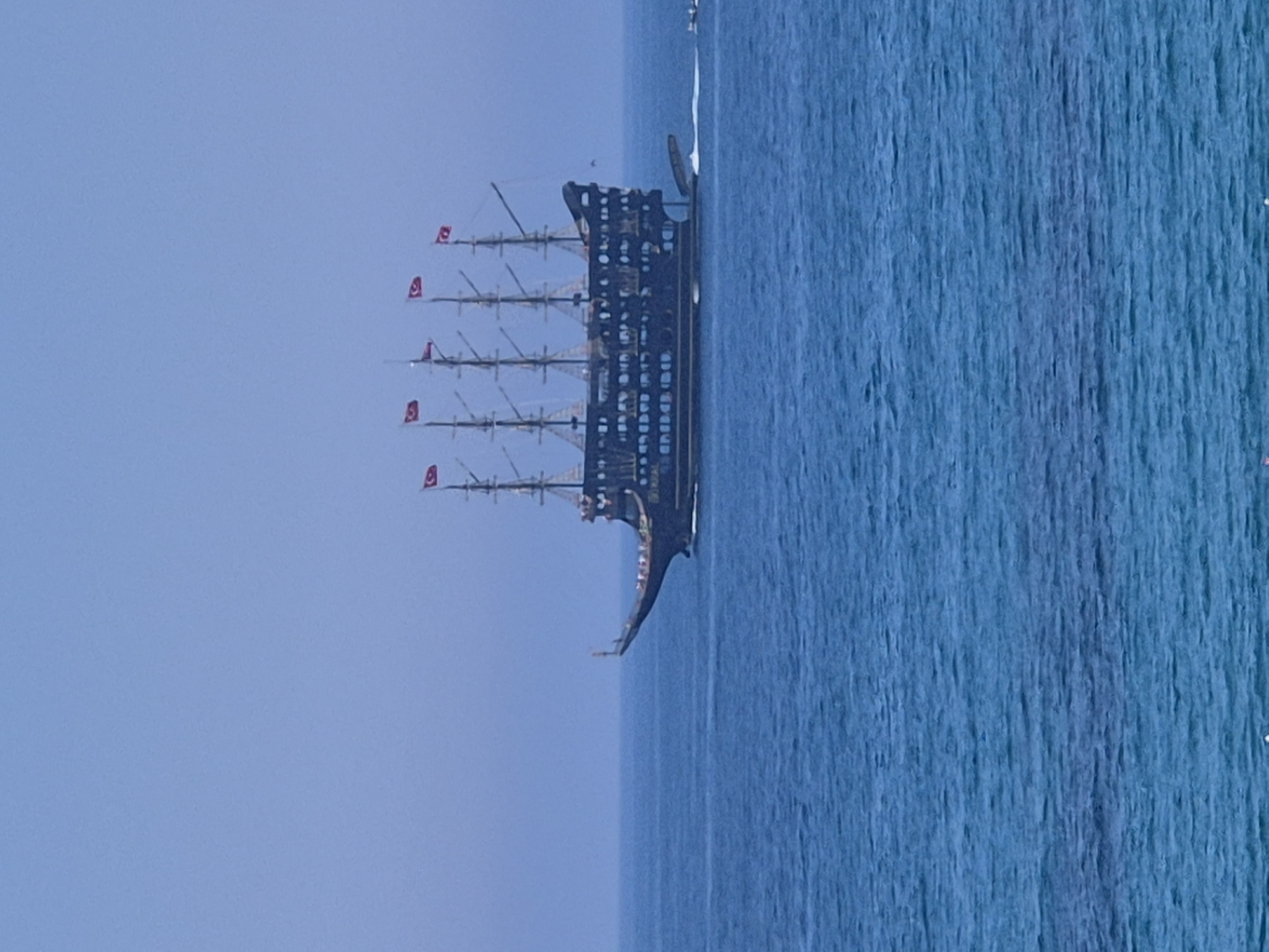 A large, dark-hulled, four-masted ship, styled as a galleon or pirate vessel, is observed underway on the deep blue waters of the Mediterranean Sea. Each of the ship's four masts flies a red Turkish flag, distinguished by a white crescent moon and star. A pronounced white wake streams from the ship's stern, indicating forward motion. Numerous small figures, presumed to be passengers, are visible on the ship's multiple decks. The sky above is a clear, uniform light blue. The scene depicts a maritime tour operation off the coast of Alanya, Turkey, a common location for such tourist excursions.
