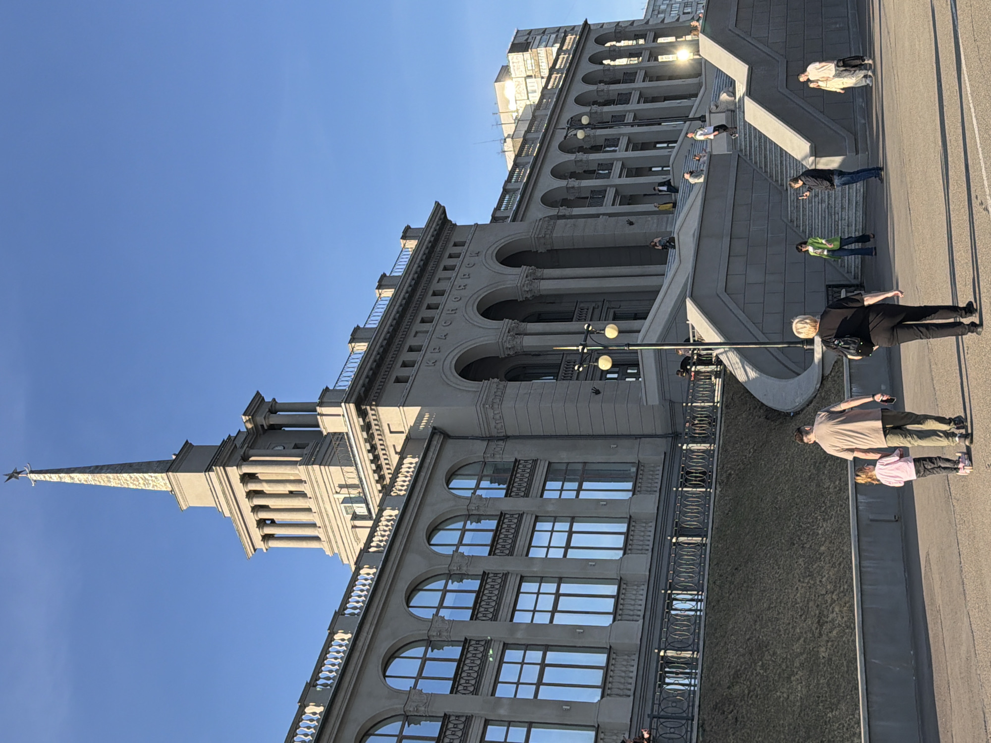 The image shows the Krasnoyarsk Krai Administration building located in Krasnoyarsk, Russia, a large, gray stone building with a pointed spire and a red star on top. The building is multi-tiered, with arched windows and decorative railings.
Several people are walking on the sidewalk in front of the building. They are casually dressed. The sky is clear and blue. A few street lights are visible.