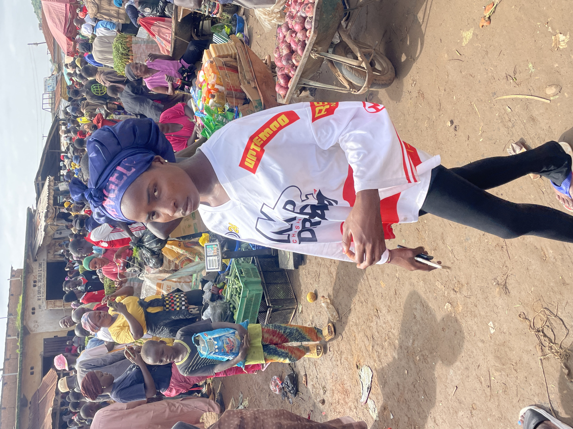 The image depicts a bustling outdoor market scene in Gura Top, Nigeria. In the foreground, a young woman wearing a blue headwrap and a white shirt with red and black lettering is walking and holding a smartphone. Her shirt features graphics that appear to be text and stylized images. She is wearing black leggings and blue and white flip-flops. Behind her, a young boy in a yellow and red tie-dye patterned outfit and yellow sandals carries a blue bag. Further back, a variety of market stalls are visible, with vendors and shoppers engaged in commerce. Several stalls display produce, including crates of cucumbers and wheelbarrows filled with onions. The ground is dirt, with scattered debris. The market is crowded with people dressed in casual clothing. Buildings with corrugated metal roofs and some concrete structures form the backdrop. The sky is overcast.