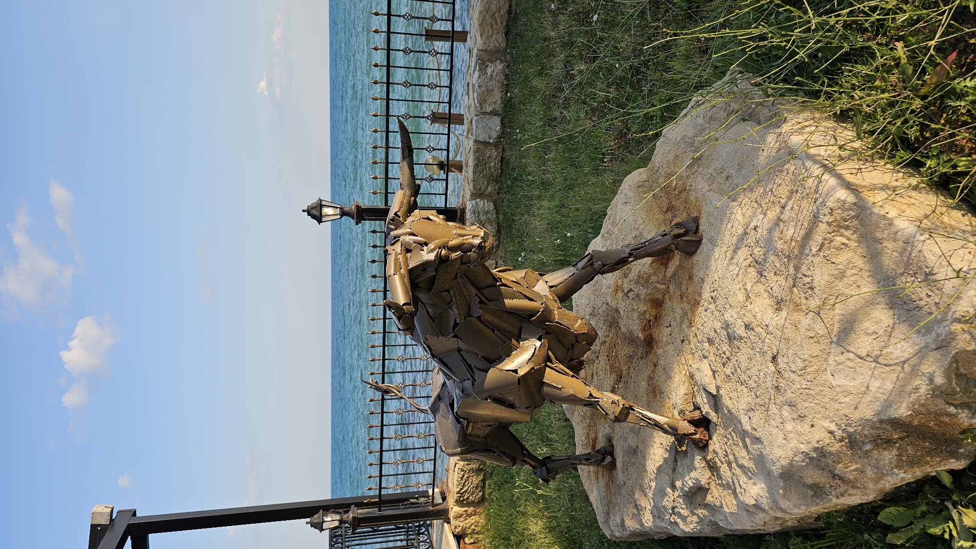 An outdoor, eye-level, medium shot captures a metal sculpture of a bull standing on a light-colored rock. The bull is made of metallic plates and is positioned in the center of the frame, facing left. Behind the bull, a black wrought-iron fence runs horizontally across the middle ground, separating the grassy area from the sea. The sea is a vibrant blue and stretches to the horizon, meeting a light blue sky with scattered white clouds. Two black lampposts with illuminated bulbs are visible; one is on the left, partially obscured by a dark structure, and another stands behind the bull, to the right of the fence. The environment suggests a coastal location, possibly within a park or a well-maintained public space. The image is taken in the city of Balchik, Bulgaria, with the Black Sea serving as the backdrop. There are no people or animals present in the scene.