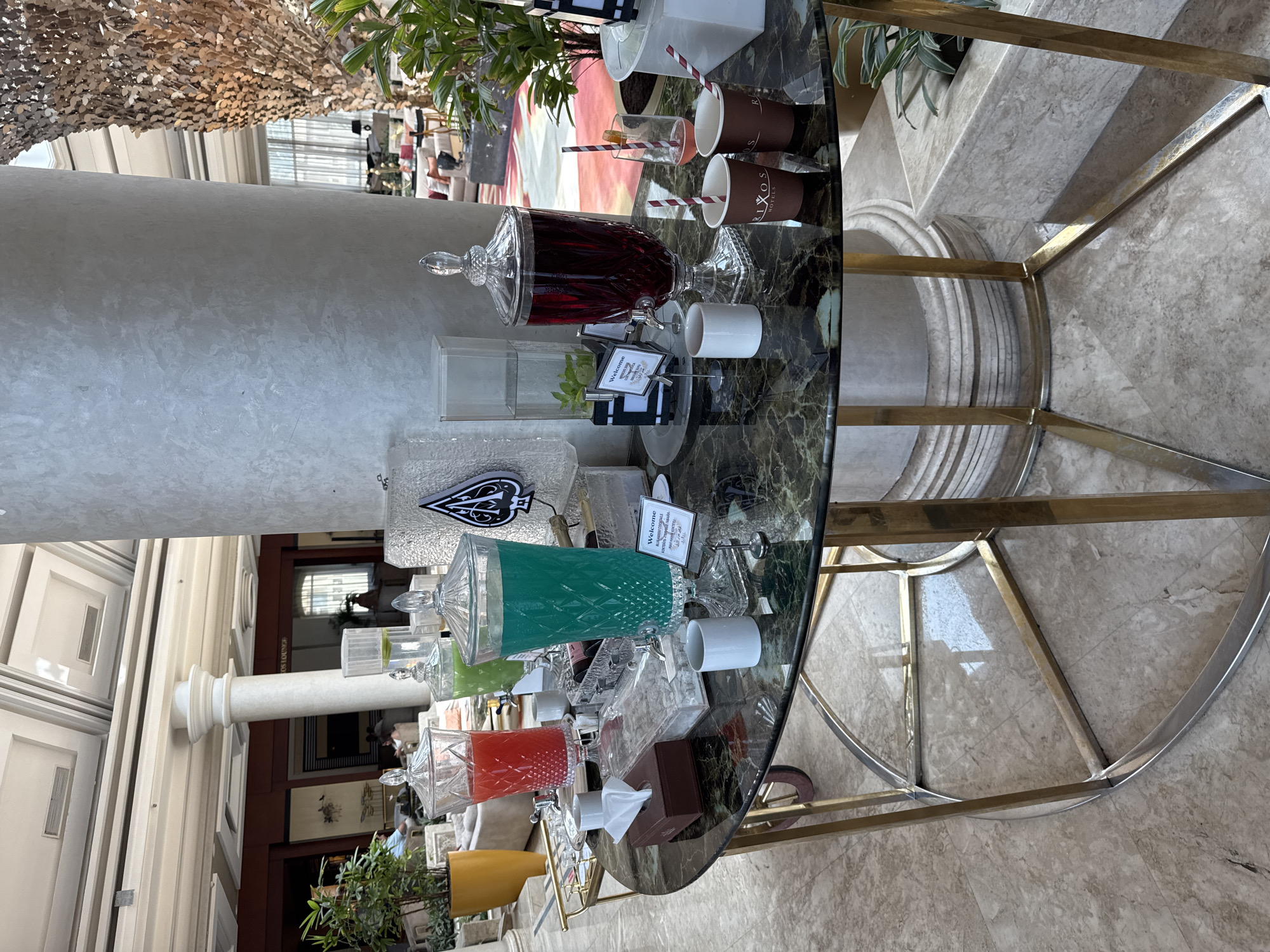 A round, dark marble table with a gold metal frame sits on a marble floor in a hotel lobby. The table is set up as a drink station, featuring several large glass dispensers filled with colorful liquids. One dispenser holds a bright red drink, another a vibrant teal, and a third contains a dark red, almost purple, beverage. To the left, a box of tissues sits next to a clear rectangular container filled with ice. Several white ceramic cups are scattered across the table, along with two brown paper cups from "RIXOS HOTELS" and two glasses with straws. A small sign with "Welcome" printed in English and Arabic is propped up near the dispensers. In the background, a large decorative pillar and lush green plants are visible, along with a glimpse of a seating area with sofas. The environment appears to be a well-lit, elegant indoor space, likely a hotel lobby in Unknown, Egypt.