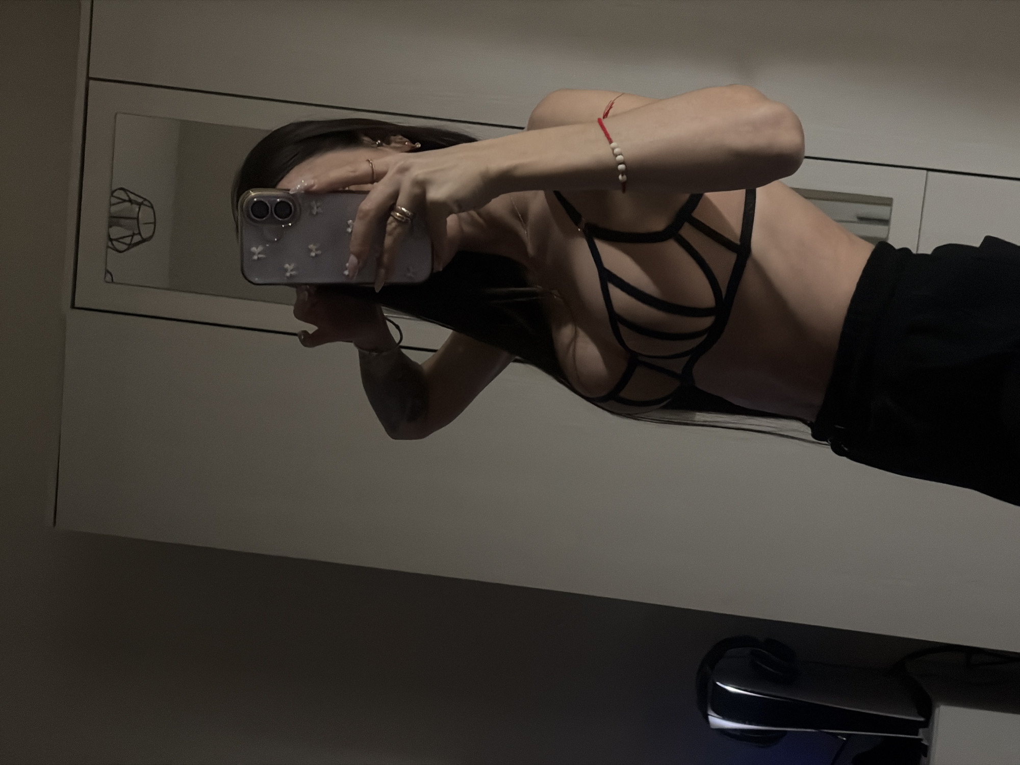 A female is taking a selfie in a mirror. She is wearing a black top and black pants. Her long dark hair is falling down her back. She is holding a phone in her right hand. She has rings on her fingers and a bracelet on her wrist. The background appears to be a room with white walls. The location is not definitively identifiable as Saint Petersburg, Russia.
