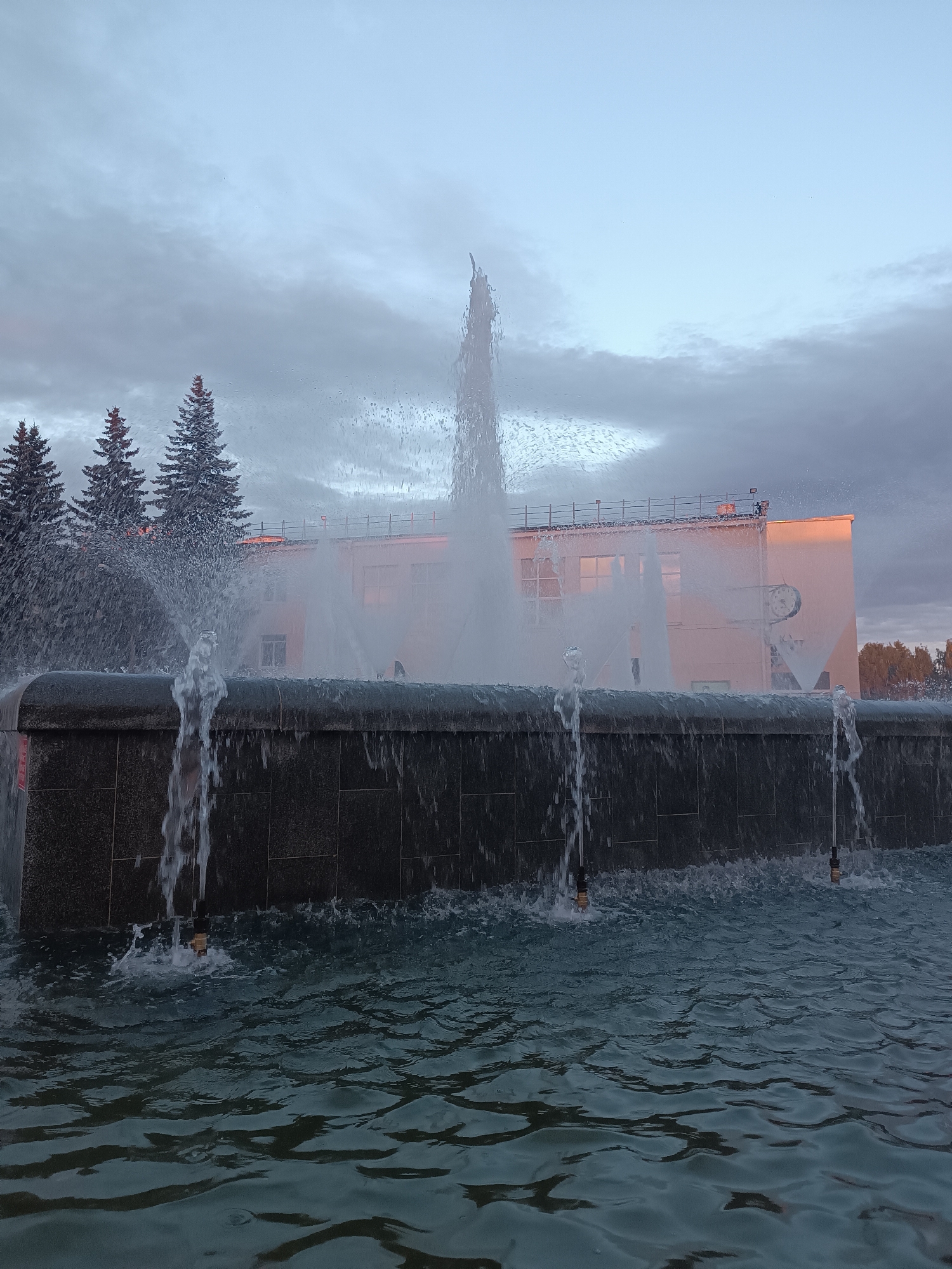 The image depicts a fountain in the city of Chaikovsky, Russia. The fountain features multiple jets of water spraying upwards and cascading over the edge of a dark granite basin. In the background, a building with windows and a light-colored facade is visible. Three tall evergreen trees stand to the left of the building. The sky is overcast with some patches of lighter blue. The water in the fountain pool is disturbed, creating ripples and reflections. The overall scene is captured during daylight, likely in the late afternoon or early evening, given the soft lighting. There are no people visible in the image.