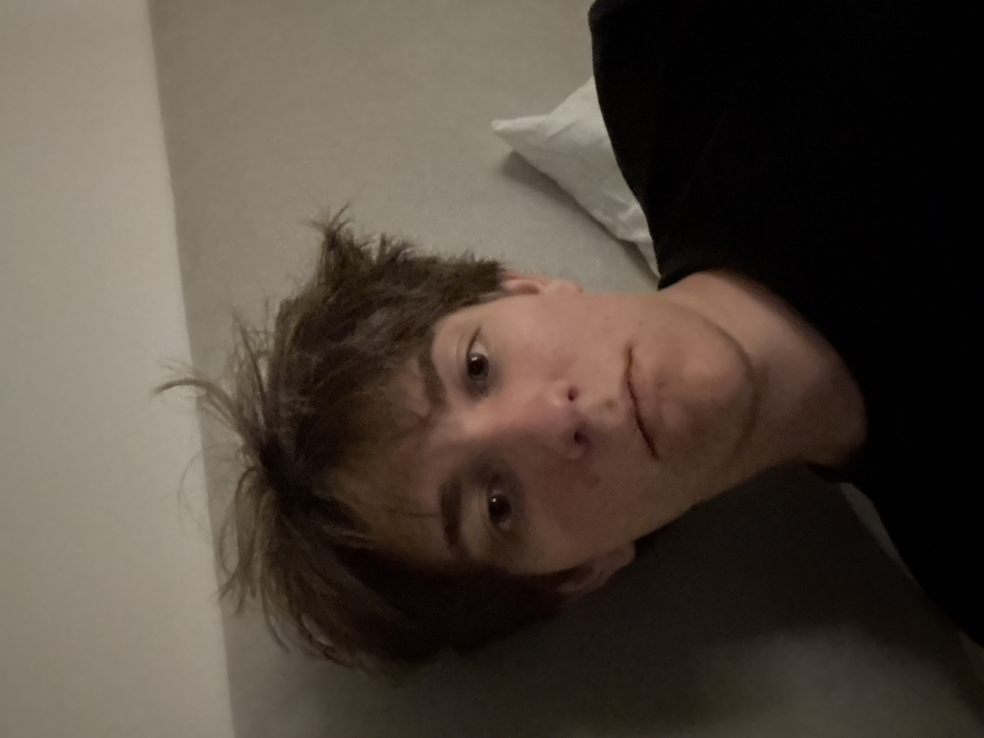 The image is a close-up, low-angle selfie of a young person with messy brown hair and fair skin, wearing a black t-shirt. The person is looking directly at the camera with a neutral expression. The background is a plain, textured wall, and a white pillow is visible to the right. The lighting is dim and appears to be coming from an overhead source, casting soft shadows. The scene is an indoor environment, possibly a bedroom or hotel room, but there are no specific elements to indicate the city of Cupra Marittima, Italy.
