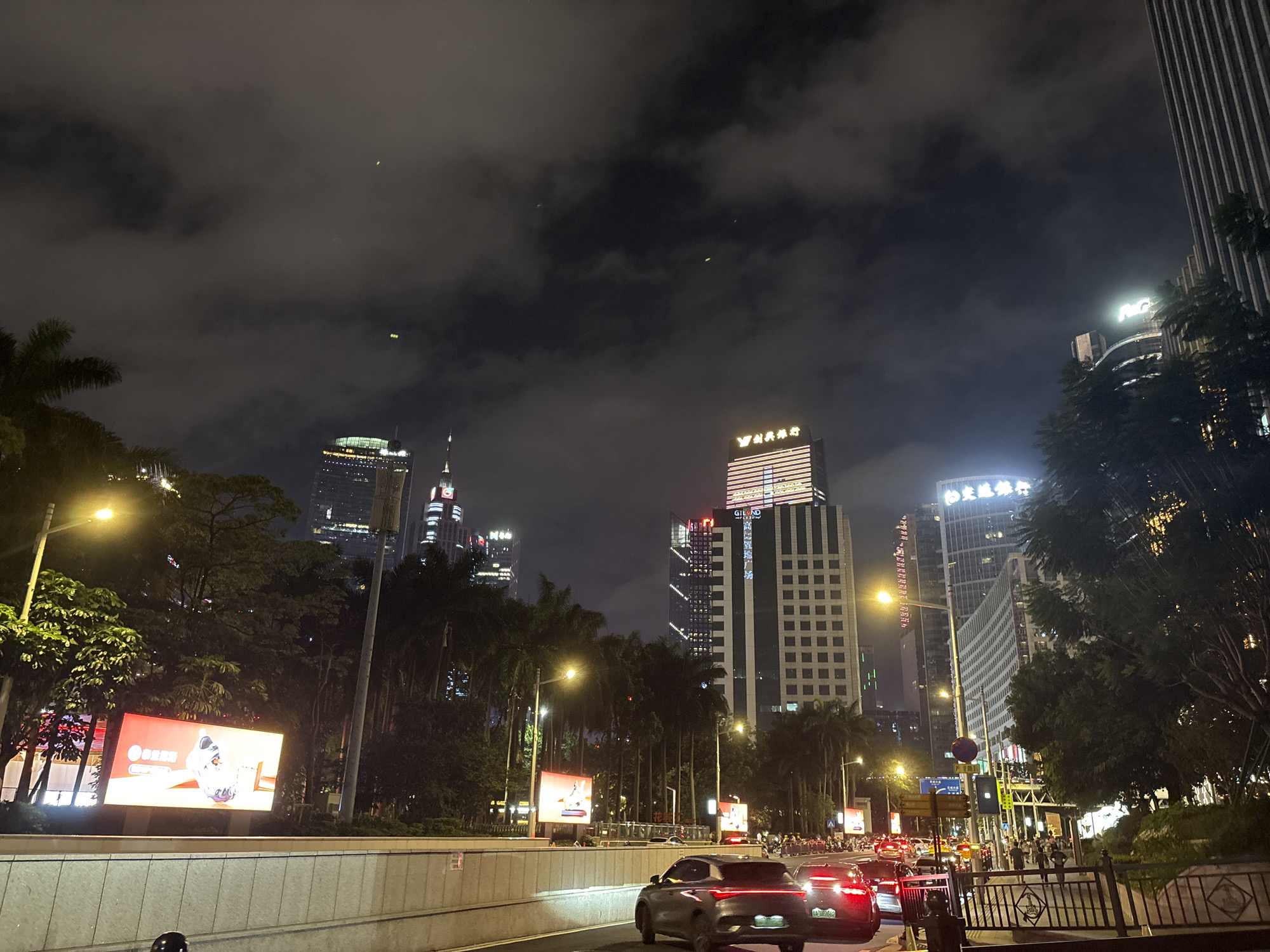 This is a nighttime, eye-level shot taken in Tianhe District, China. The scene is a busy city street with traffic, illuminated buildings, and lush greenery. The sky is dark and cloudy, with some light streaks possibly from streetlights or distant lights reflecting off the clouds.
The main subjects are the cars driving on the road, with their headlights and taillights creating streaks of light. Several tall buildings with illuminated windows and signs tower over the scene, indicating an urban environment. Among the greenery, there are palm trees, typical of tropical or subtropical regions. A large digital billboard on the left side of the image displays a product advertisement, featuring what appears to be a shoe. Another billboard further down the road also shows an advertisement.
People are visible walking on the sidewalks and crossing the street, suggesting a lively evening atmosphere. The overall mood is that of a vibrant, modern city at night, with the interplay of artificial lights and natural elements. The time of day is clearly night, and the weather appears to be overcast.
Visible text includes illuminated signage on the buildings, such as "MGM" on one of the taller buildings and Chinese characters on others, potentially indicating businesses or brand names. The digital billboards also contain text, though it is not fully legible due to the image resolution and angle. The cars are license-plated, with some legible characters and numbers.