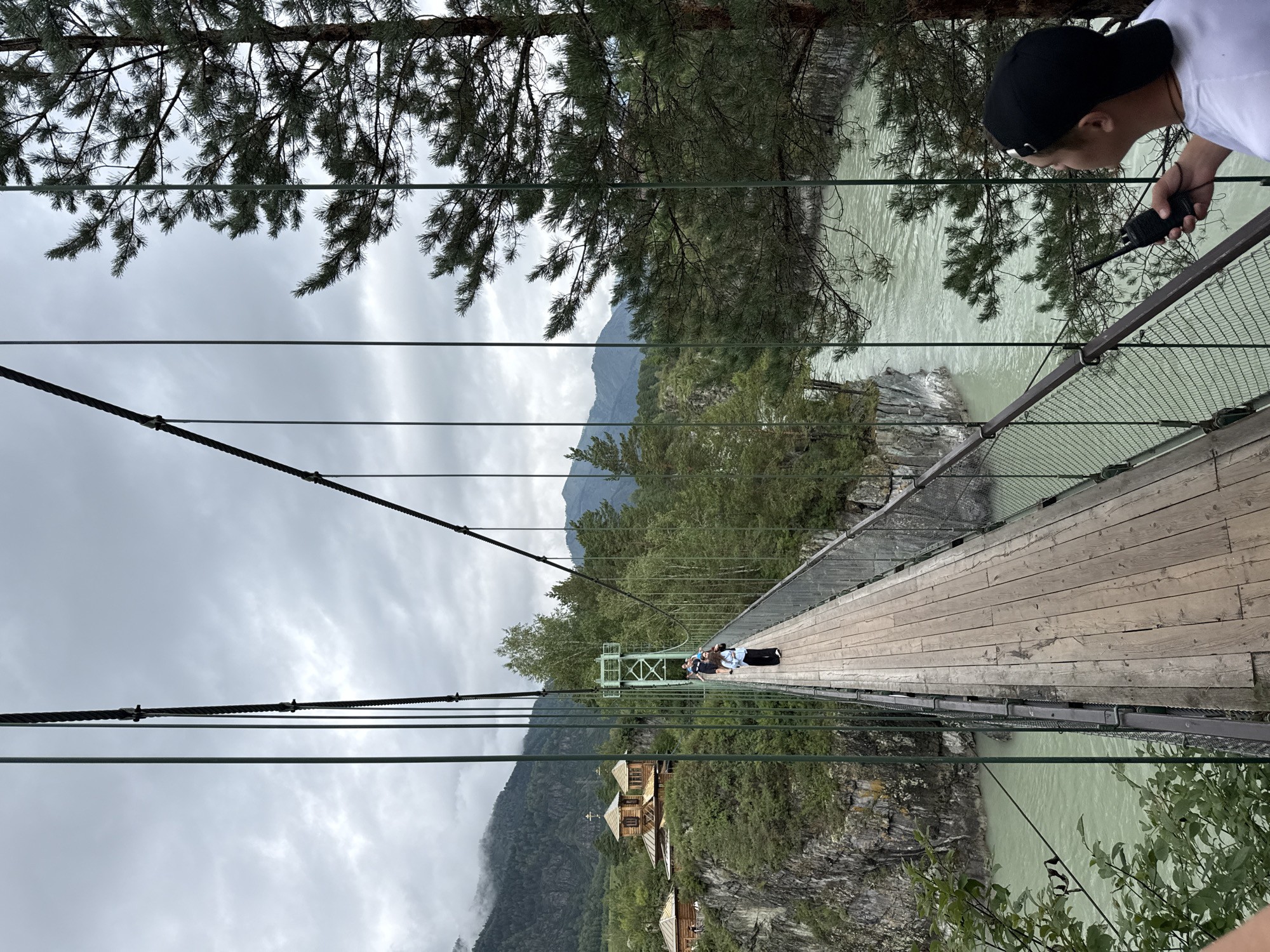 A group of people are walking across a suspension bridge spanning a jade-colored river in Chemal, Russia. The bridge has a wooden deck and metal railings. On the left side of the river, there is a cliff with a wooden building complex featuring a church with a golden dome. Lush green trees and mountains form the backdrop under a cloudy sky. In the foreground on the right side of the bridge, a person wearing a black baseball cap and a white t-shirt holds a black radio.