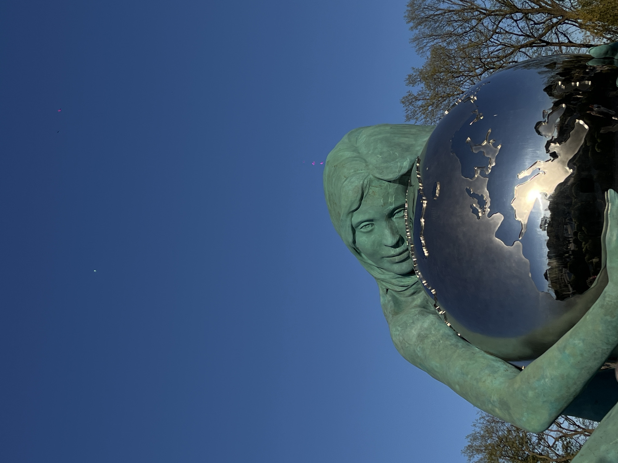 The image shows a statue of a woman holding a globe. The statue is green and appears to be made of bronze. The globe is silver and reflective. The background is a clear blue sky with some trees visible in the upper right corner. The statue is located in the city of Krasnodar, Russia.