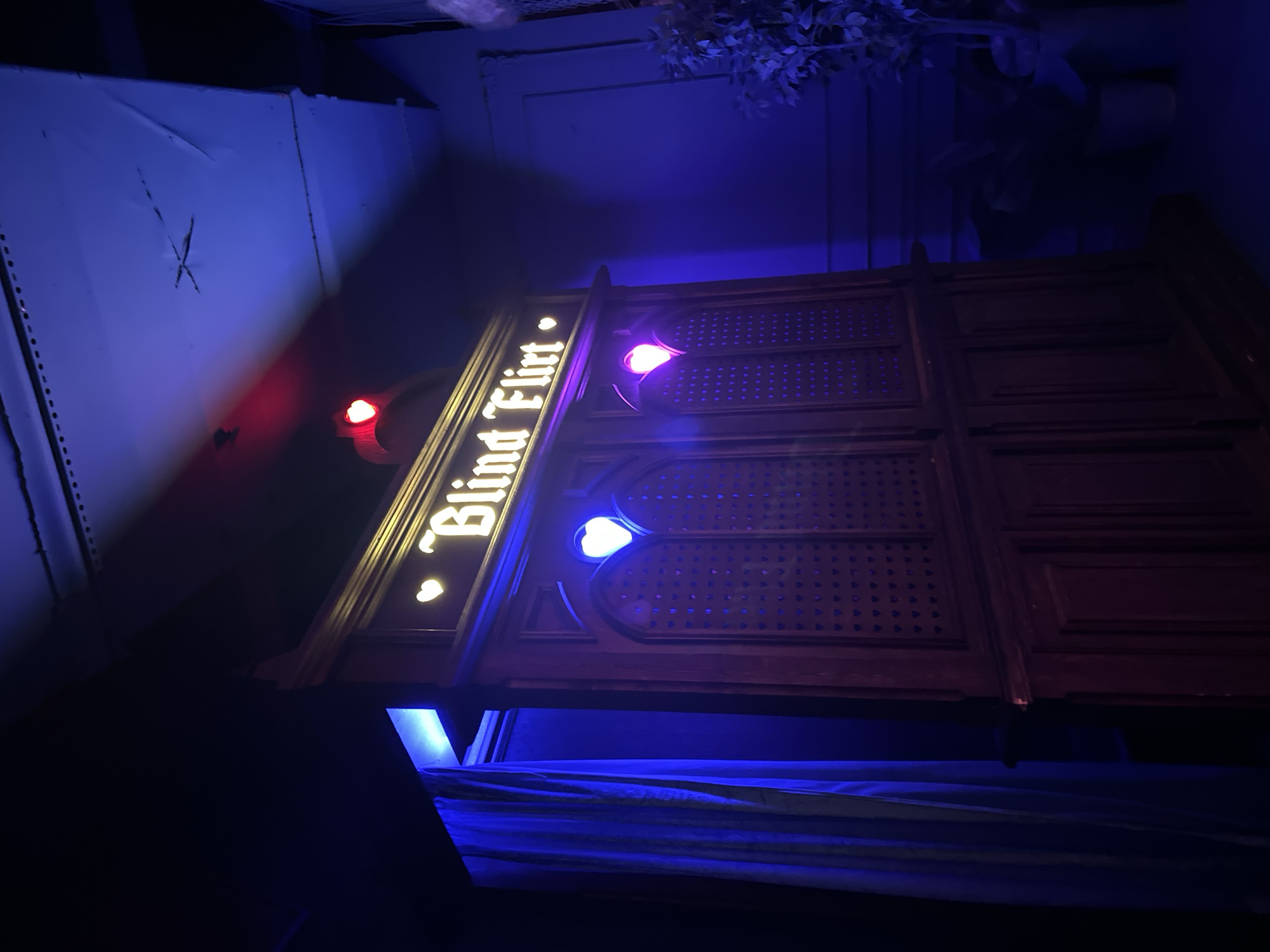 The image shows a sign that reads "Blind Flirt" illuminated with heart-shaped lights. The sign appears to be made of dark wood. The background is dark, illuminated with blue and red lights. A curtain is visible below the sign. The scene appears to be indoors, possibly in a bar or club. There is no mention of location in city of Sofia, Bulgaria.