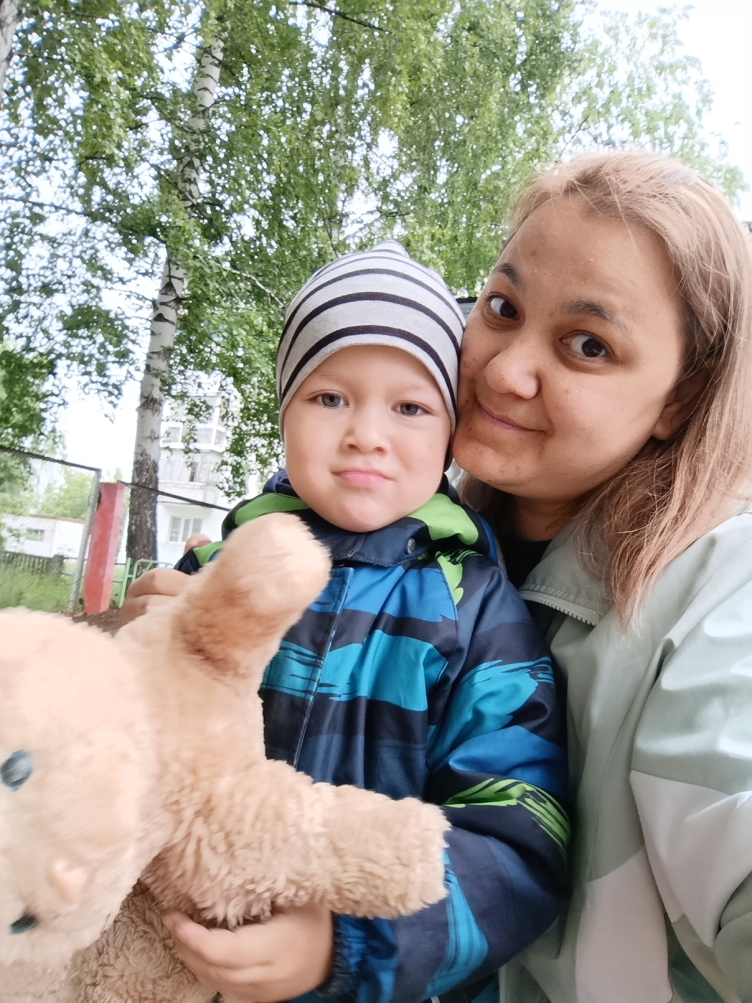 A close-up outdoor selfie shows a woman and a young boy, likely a mother and son, in Chaikovsky, Russia. The woman, with light-colored hair and a faint smile, leans her head towards the boy, looking directly at the camera. She is wearing a light mint green or grey jacket. The boy, positioned to her left, wears a grey and black striped beanie and a blue, green, and black patterned winter jacket. He has a neutral to slightly curious expression, also looking towards the camera. He is holding a light brown, fluffy teddy bear with blue eyes, which is partially visible in the foreground.
The background features lush green foliage, suggesting an outdoor setting during a warm season. A prominent white-barked birch tree trunk is visible behind them, along with other trees. In the mid-distance to the left, a metal fence and parts of a residential building with windows can be seen. The lighting appears bright and natural, indicating daytime, possibly with an overcast sky as there are no harsh shadows. The overall impression is a casual, affectionate moment captured outdoors. No visible text is present in the image.