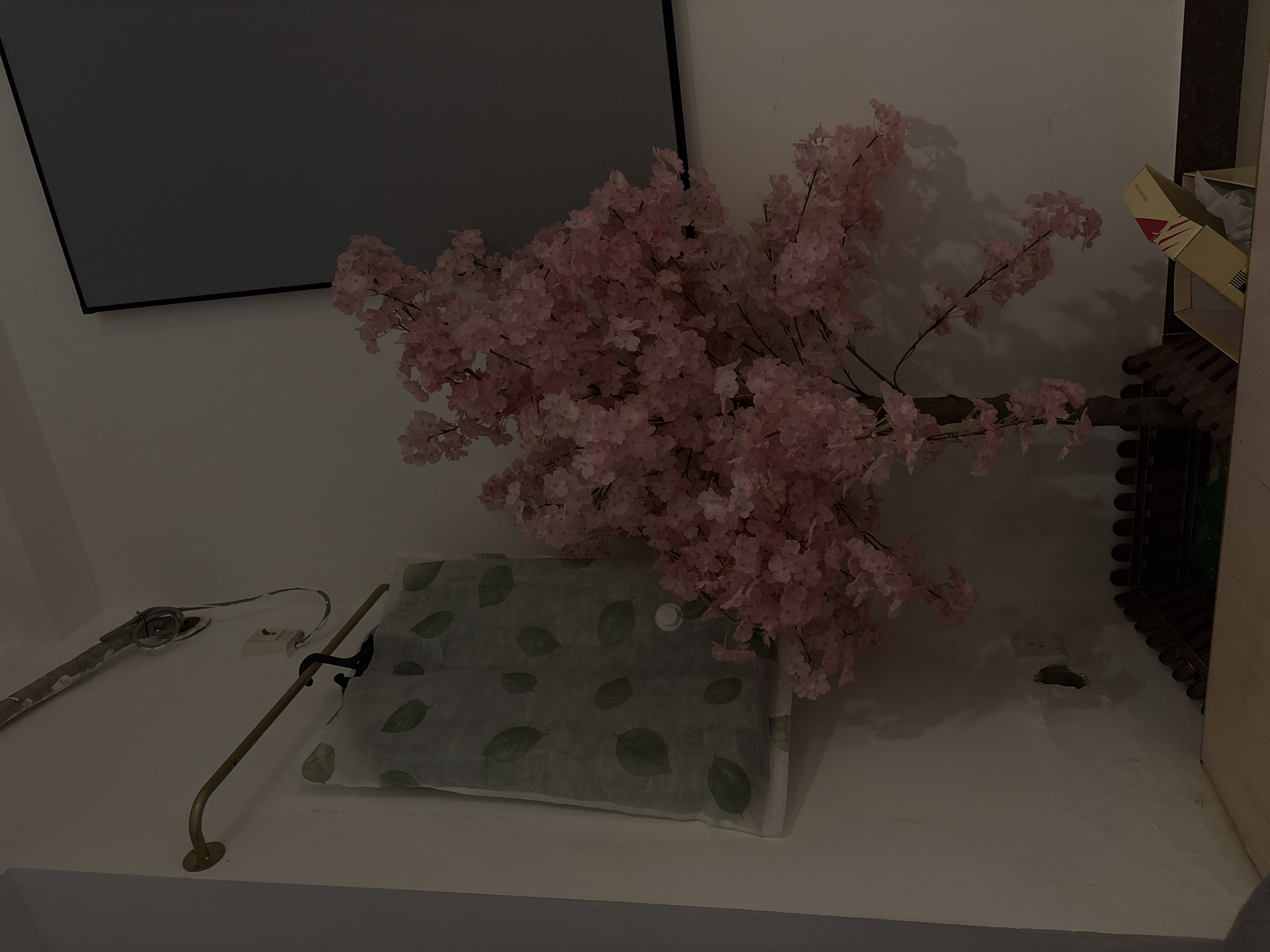 The image depicts a still life arrangement on a white shelf. Dominating the scene is a large branch of artificial pink cherry blossoms, their delicate petals clustered densely. To the left of the blossoms, a large, flat object covered in a patterned green material with leaf motifs is visible. A metal rail, possibly a handrail or part of a display fixture, curves across the shelf in front of the object and the blossoms, anchored by a circular base. To the far left, a grey cylindrical object, perhaps a tool or a piece of equipment, is also present, connected by a wire to a small white electrical outlet on the wall. In the background on the right, a brown wooden structure with a stacked, tiered appearance is partially visible, alongside an open cardboard box with some white paper inside. The wall behind the objects is white, with subtle shadows cast by the cherry blossom branches. The lighting appears to be artificial and dim, creating a somewhat moody atmosphere. The scene suggests an indoor setting, possibly a home or a retail display area. No people are visible, and the overall impression is one of decorative arrangement rather than an active event. The time of day is indeterminate due to the lighting. No discernible text is present in the image.
