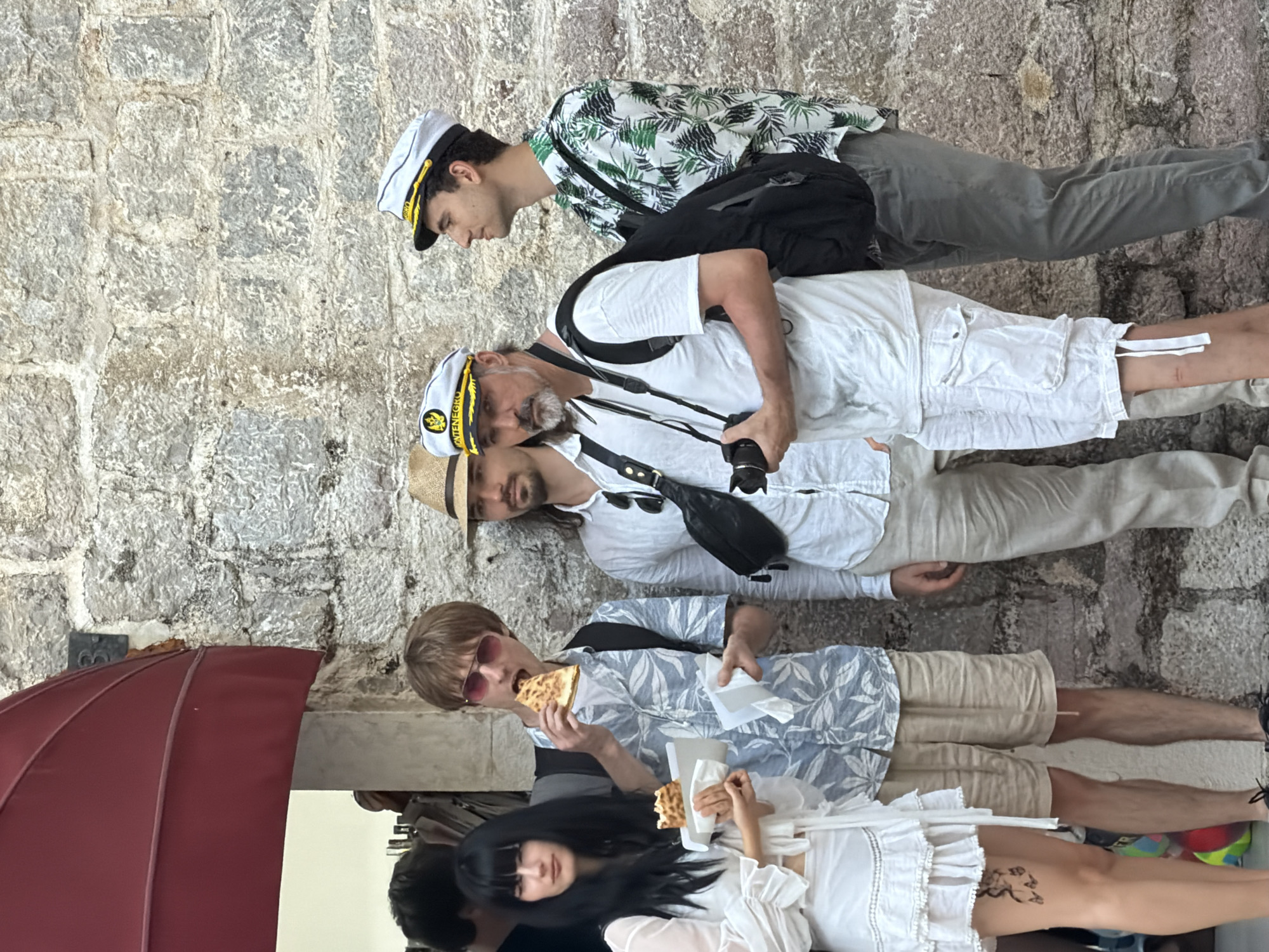 A group of five individuals are gathered against a textured stone wall in Budva, Montenegro. From left to right, the first individual is a woman with long black hair, wearing a white ruffled dress and holding a piece of food. Next to her, a man with short brown hair and pink sunglasses is eating a slice of pizza. He is wearing a light blue and white patterned short-sleeved shirt and beige shorts. To his right, a man with dark hair and a straw hat is wearing a white shirt and beige pants. The fourth individual, a man with a gray beard, is wearing a white t-shirt, white cargo shorts, and a white captain's hat. He is holding a camera. The fifth person, a man with dark hair, is wearing a white captain's hat and a green and white palm-print short-sleeved shirt, paired with gray pants. A large red awning is visible in the upper left corner of the image. The ground appears to be paved, and in the lower left, a colorful beach ball is partially visible.