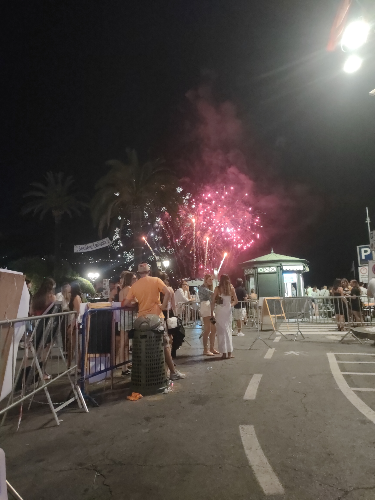 A nighttime photograph captures a fireworks display over a waterfront area, likely in Rapallo, Italy. In the foreground, a street is marked with white lines. Several people are gathered, observing the fireworks. Some individuals are standing behind metal barriers, while others are conversing.
A prominent feature is a green, octagonal kiosk with a pointed roof, situated in the midground. Behind the kiosk, the fireworks explode in shades of pink and red against the dark night sky. Palm trees with decorative lights are visible in the background, illuminated by the fireworks. A banner reading "Sestriere Cerisola" hangs between two palm trees.
The scene suggests a celebratory event, with people dressed in casual summer attire. The ground appears to be paved, possibly asphalt, and there are trash receptacles and various temporary barriers present. The overall atmosphere is festive and crowded.