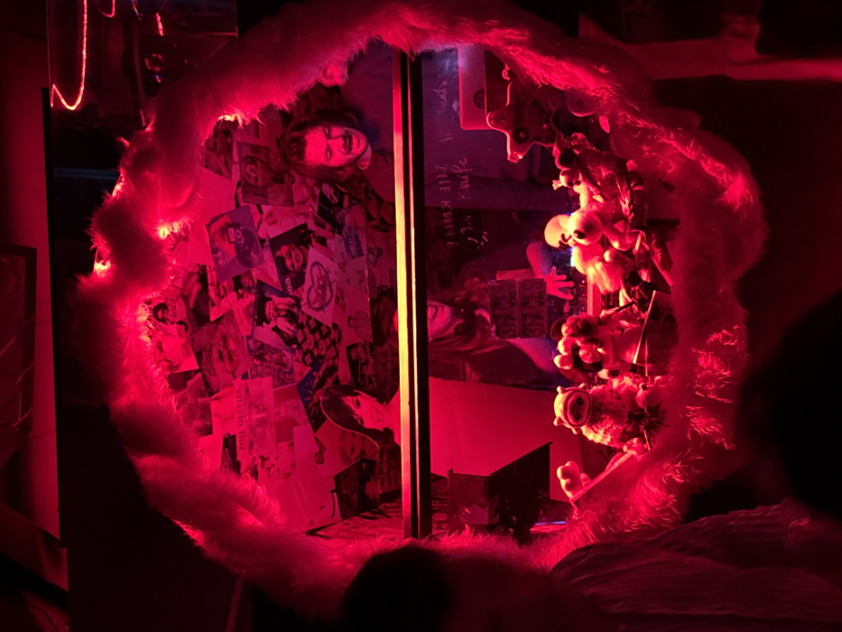 The image depicts an interior scene, likely a bar or music venue, characterized by deep red ambient lighting. The primary view is framed through an irregular, fluffy, white or light-colored border, possibly faux fur, which occupies a significant portion of the foreground on the left and right, partially obscuring a blurred person on the far left.
Within this frame, the scene is divided horizontally. The upper section features a wall adorned with a dense collage of various printed images, including album covers, magazine cutouts, and photographs. Among these, a female cutout with dark hair and bangs is visible on the left, and a man with long, wavy dark hair and a mustache is visible on the right, looking forward.
Below this collage, a transparent barrier or glass partition separates the upper background from the lower area. On the glass, reflections show text in reverse, reading "THE KNIFE" and "hi cats!", indicating elements located behind the viewpoint. A person, partially obscured, is reflected, appearing to operate a DJ mixer or similar electronic music equipment with both hands, positioned behind a laptop also reflected on the surface.
Directly on the surface behind the glass, and in front of the reflected person, a collection of plush toys is arranged. These include a light-colored pig-like figure in the center, surrounded by other diverse stuffed animals and figures, all illuminated by the strong red light.
Based on the distinct aesthetic, including the furry frame, plush toy display, red lighting, and music-oriented setup, the location is identified as **PopUp! du Label, situated in the 12th arrondissement of Paris, France.** The scene conveys an eclectic, nocturnal atmosphere characteristic of a performance or social venue.