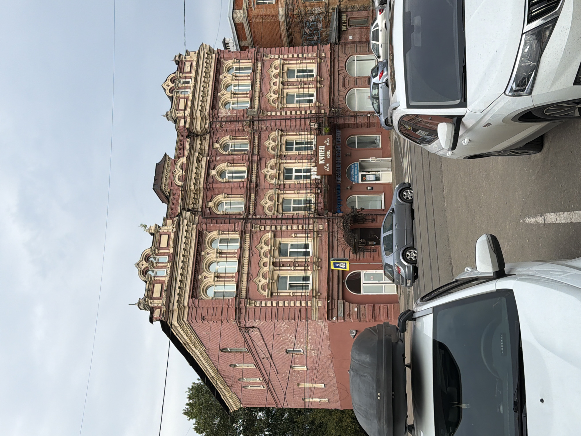 This is a medium-angle, eye-level, vertical outdoor photograph taken on a cloudy day in Nizhny Novgorod, Russia. The main subject is a historic, multi-story brick building with ornate architectural details. The building is painted a muted pink and features cream-colored decorative elements around the windows and along the roofline. The windows on the upper floors are arched, while those on the ground floor are rectangular. A sign above the main entrance reads "КУПЕЦ HOTEL" (Kupez Hotel) in Russian.
In the foreground, the top of two white cars is visible. The car on the left has a large black cargo box mounted on its roof. To the right of the main building, a portion of another brick building, darker in color, is visible. Yellow pedestrian crossing signs and a ladder on the side of the main building are also present. Several other vehicles, including silver sedans, are parked or driving in the street. Electrical wires are strung across the facade of the building. The scene depicts a streetscape in a city environment.
