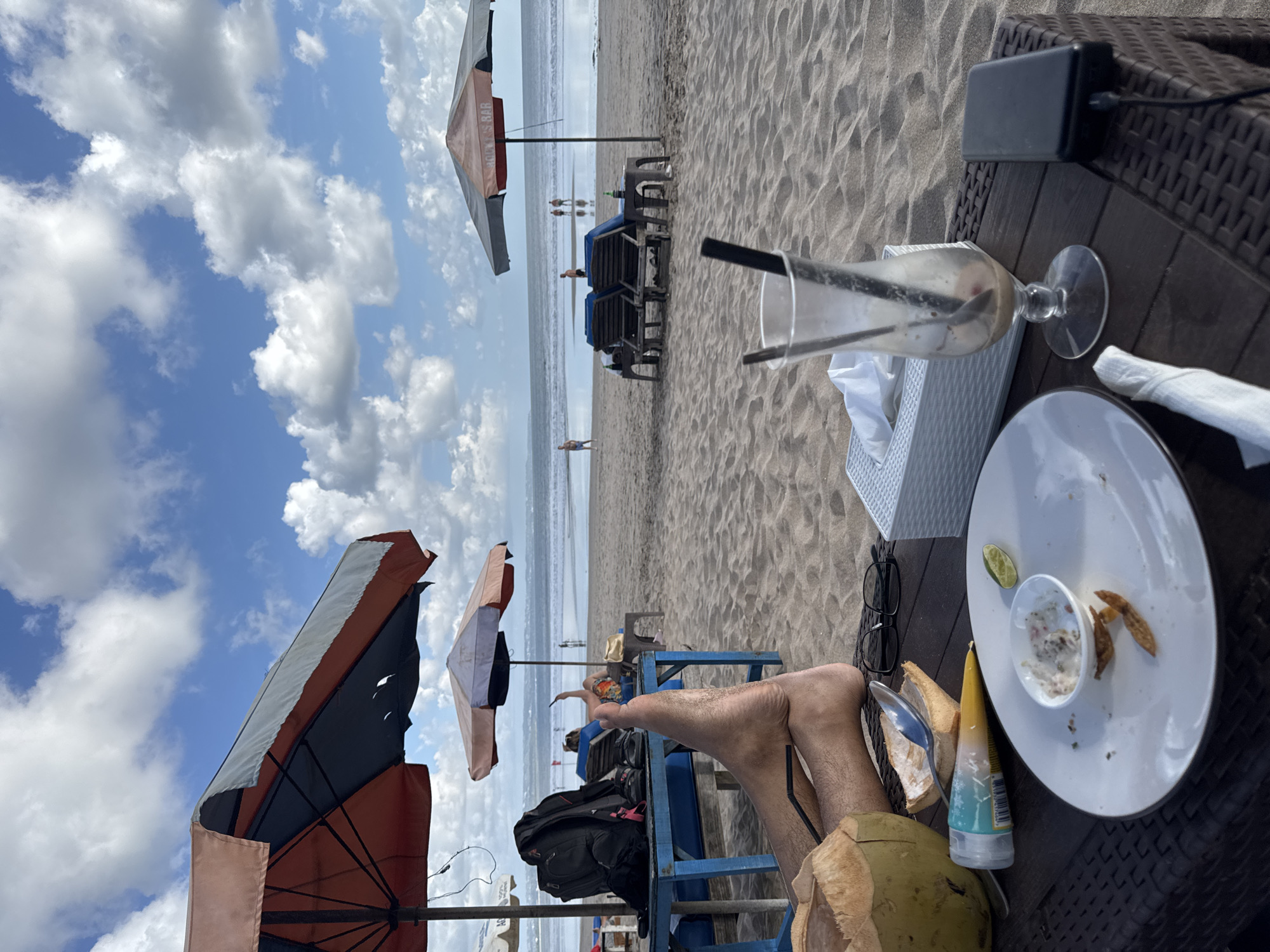 This is a medium shot of a beach in Kuta, Indonesia, on a bright, cloudy day. In the foreground, a brown wicker table is laden with remnants of a meal and drinks. A half-eaten coconut with straws sits to the left of the frame, next to a used plate with a lime wedge and some food scraps. A nearly empty hurricane glass with straws and a spoon stands to the right, beside a white tissue box and a black power bank connected to a cable. A pair of black-rimmed glasses lies on the table near a tube of what appears to be sunscreen.
In the mid-ground, the sandy beach stretches towards the ocean, dotted with blue lounge chairs under large, colorful umbrellas. A person's legs are visible resting on a blue lounge chair in the left mid-ground, with a black backpack placed on a blue table behind them. Several people are seen walking on the beach and wading in the shallow water in the distance. The ocean is calm, with gentle waves rolling towards the shore. The sky is a vibrant blue, filled with fluffy white clouds.
