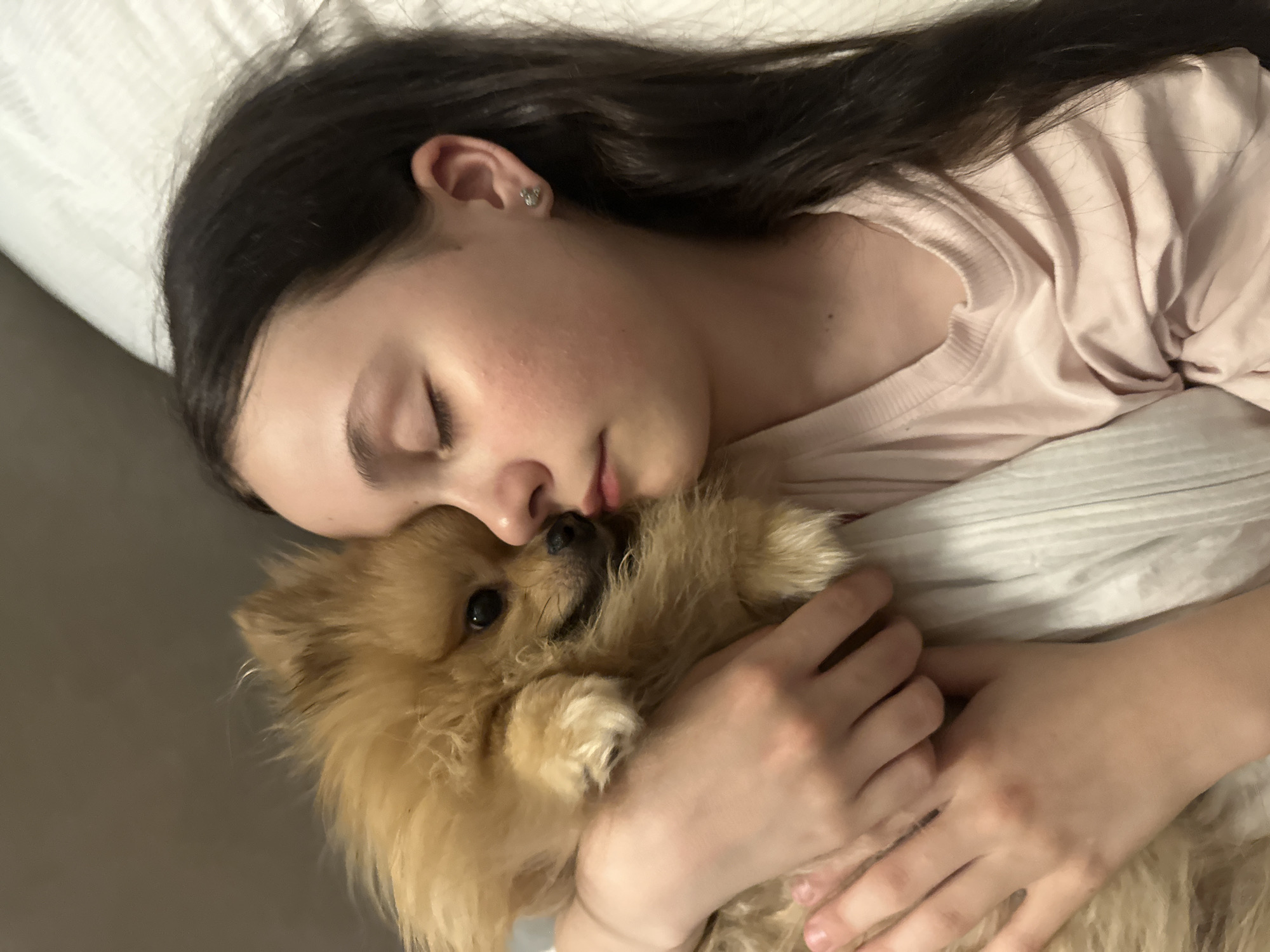 A young woman with long, dark hair is sleeping in a bed with a Pomeranian dog. She is wearing a light pink shirt. She has fair skin, closed eyes, and a small stud earring in her left ear. Her face is pressed against the dog's fur, and her hands are wrapped around it. The dog is facing the viewer and appears calm. The background consists of white bedding. It is not possible to tell the location in the city of Votkinsk, Russia from this image.