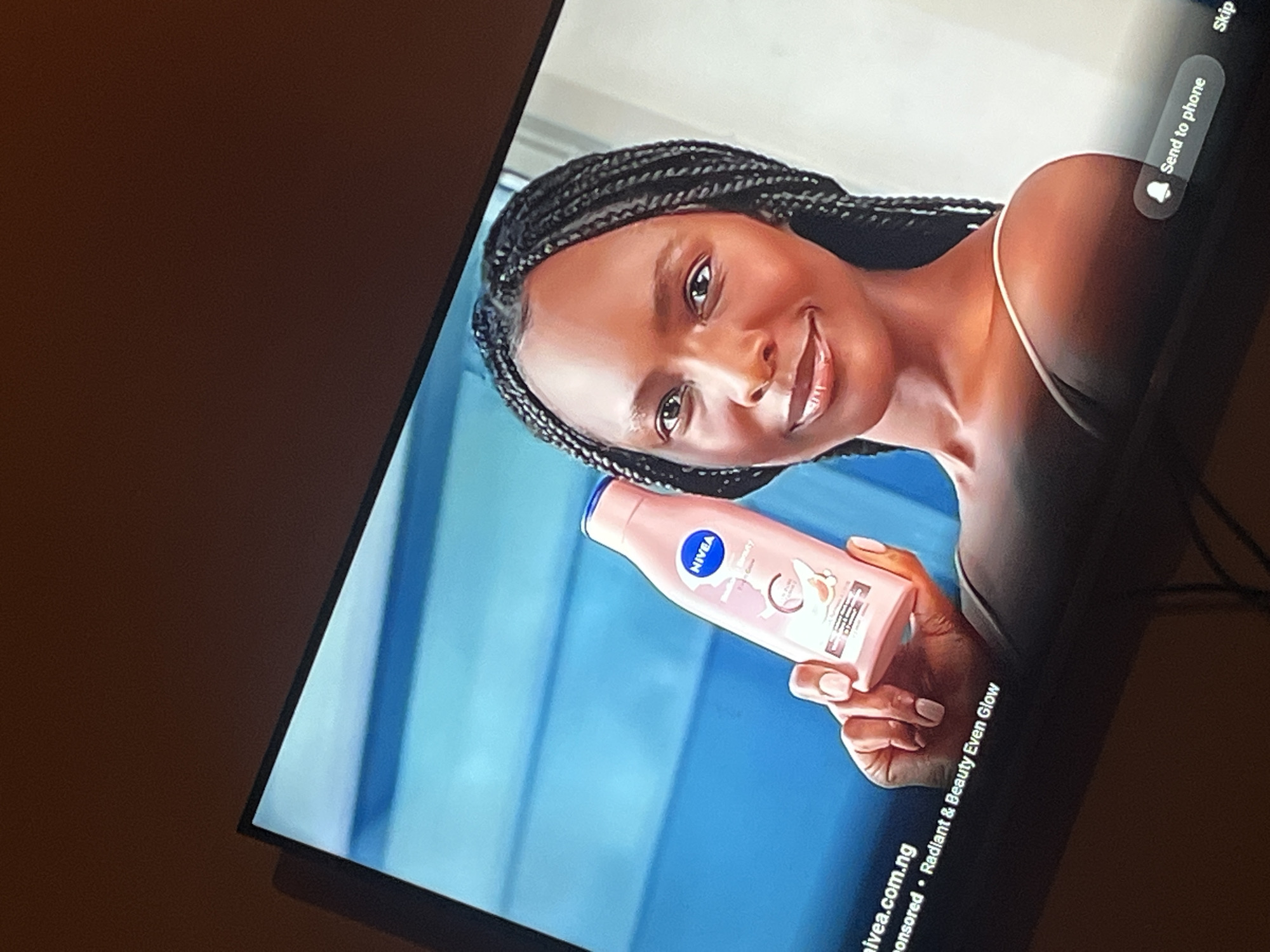 The image shows a close-up of a woman's face and upper body as she holds a bottle of Nivea body lotion. She has dark skin and her hair is styled in braids. She is smiling and looking towards the viewer. The body lotion bottle is pink and has the Nivea logo on it. The background is a bright, solid blue. Text at the bottom of the screen reads "nivea.com.ng Sponsored - Radiant & Beauty Even Glow" and a button with a bell icon says "Send to phone". The image appears to be a screenshot from a digital advertisement or video being displayed on a television screen. There is no indication that the scene is located in Gura Top, Nigeria or any other specific city.