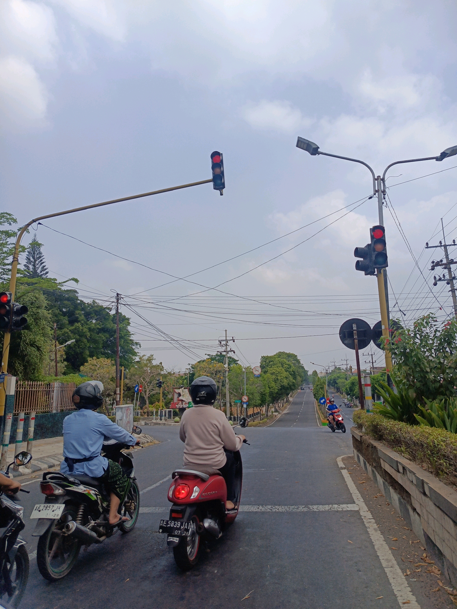 The image depicts a street scene in Sukowinangun, Indonesia, on an overcast day. Two individuals are riding motorcycles, facing away from the camera and proceeding down a paved road. The rider on the left wears a light blue shirt and a dark helmet, operating a motorcycle with a white license plate reading "AE 2857 JK". The rider on the right wears a light brown long-sleeved shirt and a dark helmet, riding a red scooter with a license plate that reads "AE 5839 JAI". Several traffic lights and street lamps are visible along the road. The road is lined with trees and vegetation on both sides, with a low stone wall on the right. Overhead, a complex network of power lines crisscrosses the sky. In the distance, more trees and buildings are visible, along with other vehicles on the road. The sky is mostly covered by clouds, with patches of lighter sky visible.