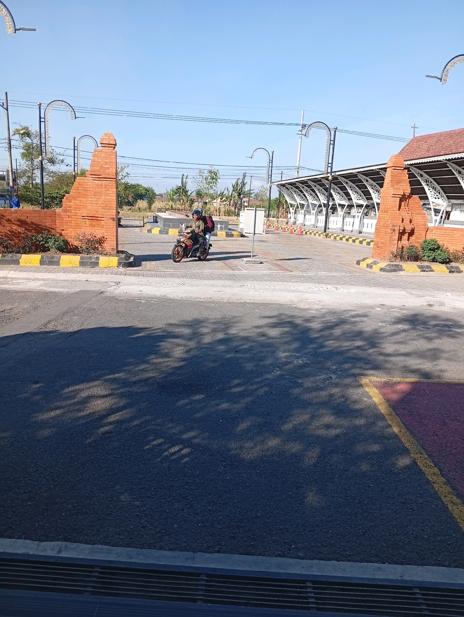 A motorcyclist wearing a helmet and a backpack rides a motorcycle through an open brick gateway under a clear blue sky in Tebon, Indonesia. The gateway, constructed from reddish-brown bricks, features decorative arches and pillars. Beyond the gateway, a long, covered structure with a white, patterned roof supported by white, ornate beams is visible. The ground in front of this structure is paved with interlocking bricks and marked with yellow and black safety lines. The foreground is an asphalt road with a faint pattern of shadows cast by trees. The scene appears to be an entrance to a public area or a facility.