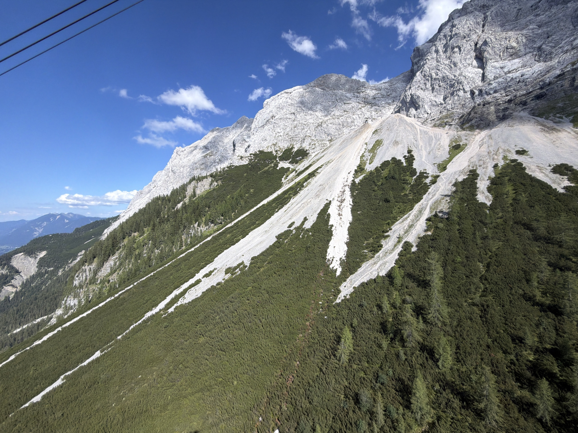 This media file is an outdoor, high-angle shot of a mountainous landscape, possibly taken from a cable car given the presence of thick cables in the upper left corner. The scene is dominated by a steep, rocky mountain face on the right, characterized by stark grey stone with visible strata and patches of scree. The lower slopes are covered in dense, dark green coniferous trees, giving way to lighter green vegetation and rocky paths further up. White streaks, likely scree or snowmelt paths, run down the mountainside through the trees and rock. In the distance, to the left, are further mountain ranges under a bright blue sky dotted with fluffy white clouds.
The time of day appears to be daytime, with strong sunlight casting shadows and highlighting the textures of the rock and foliage. The weather seems clear and fair, suitable for outdoor activities. The location is likely a mountainous region in Germany, specifically Grainau, known for its alpine scenery. No people, animals, or text are visible in the image. The overall impression is one of natural grandeur and a remote, rugged environment.