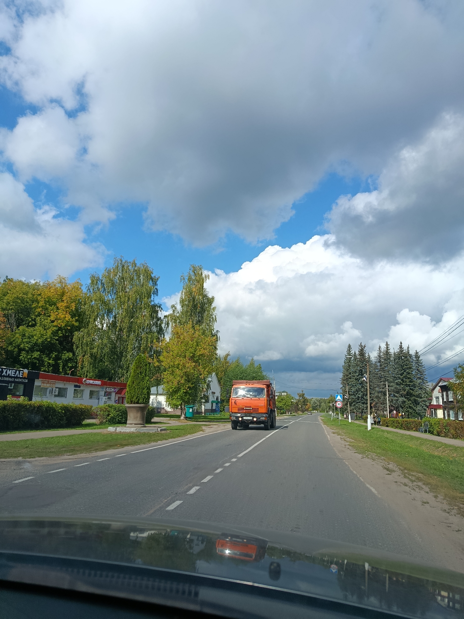 The image shows a view from a vehicle traveling on a road in Verea, Russia. An orange truck is driving on the road. To the left of the road, there are buildings, trees, and a large planter. To the right of the road, there are more trees and houses. The sky is filled with clouds.