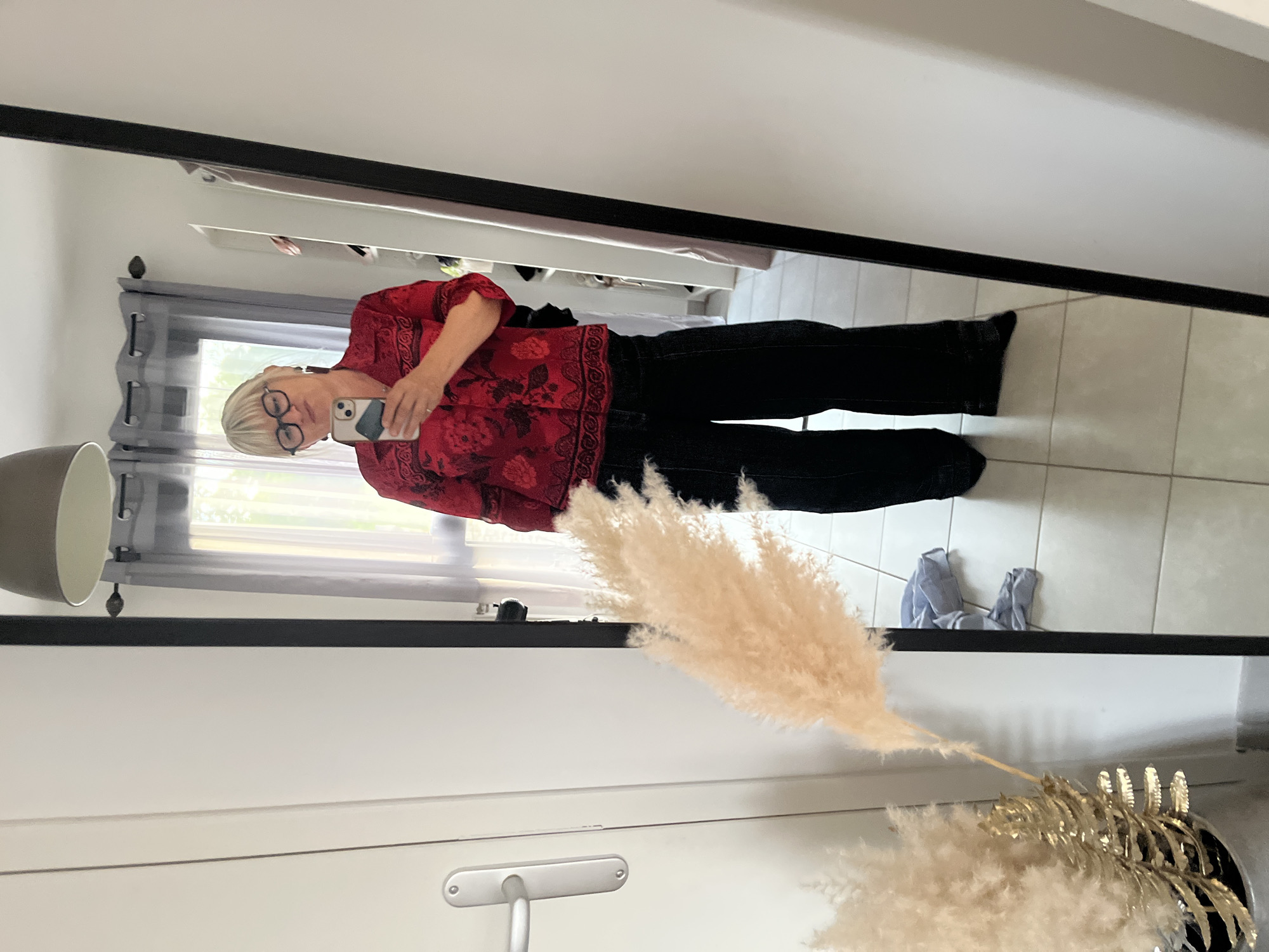 A woman is visible in a mirror reflection, holding a smartphone, wearing glasses, a red blouse with floral patterns, and dark pants. She is standing in a room that appears to be indoors. A lamp, window with curtains, and a door are visible in the background. A vase with decorative plumes is in the foreground. The location is Chambœuf, France.