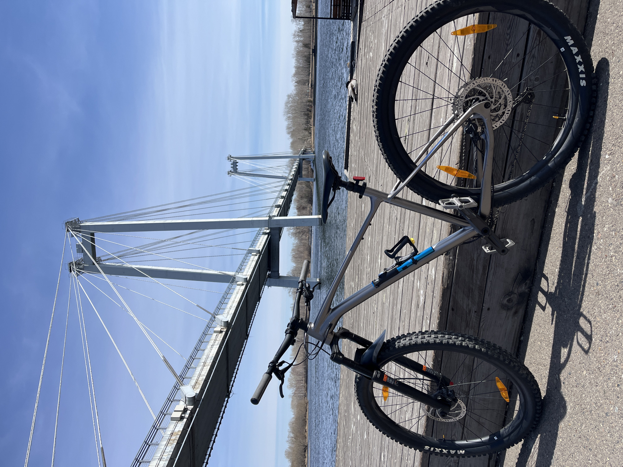 A bicycle is lying on its side on a wooden dock. In the background is the Vinogradovsky Bridge over the Yenisey River in Krasnoyarsk, Russia. The sky is blue with a few clouds. The water is choppy.