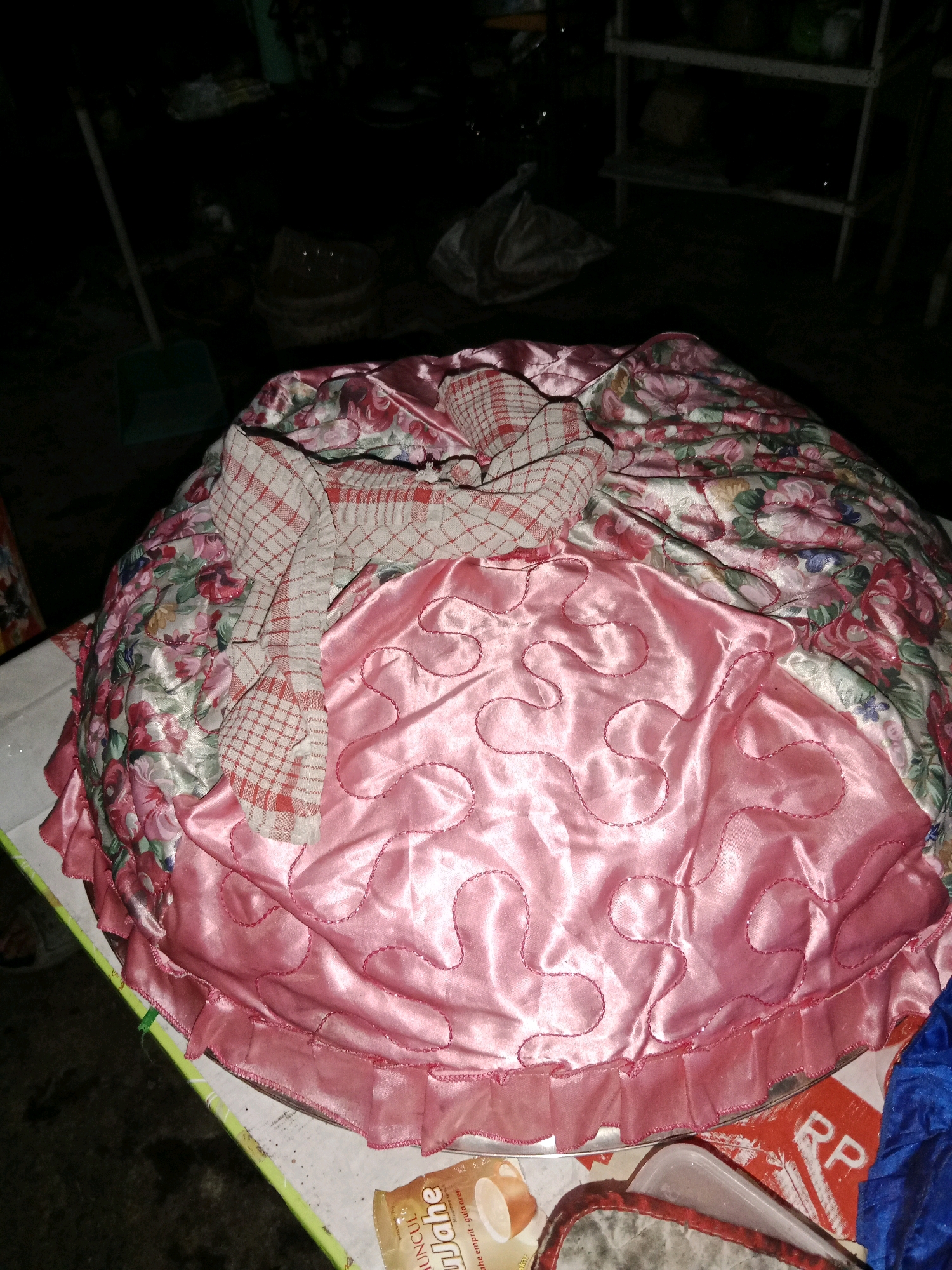 A large, round object covered with fabric is the central focus of this image. The fabric has a floral pattern in shades of pink, red, and green, with a sheen that suggests it is satin or a similar material. Over this, a piece of checkered fabric, predominantly red and white, is draped. The bottom portion of the object is covered by a solid pink fabric with a ruffled edge, also with a sheen. The object appears to be resting on a tray or a flat surface. In the background, the scene is dark and somewhat cluttered, with a broom leaning against a wall on the left and shelves with various items on the right. A portion of a red and white label with the letters "RP" is visible at the bottom right, suggesting a product or packaging. The overall lighting suggests an indoor or outdoor evening setting. The location is identified as Ringinrejo, Indonesia.
