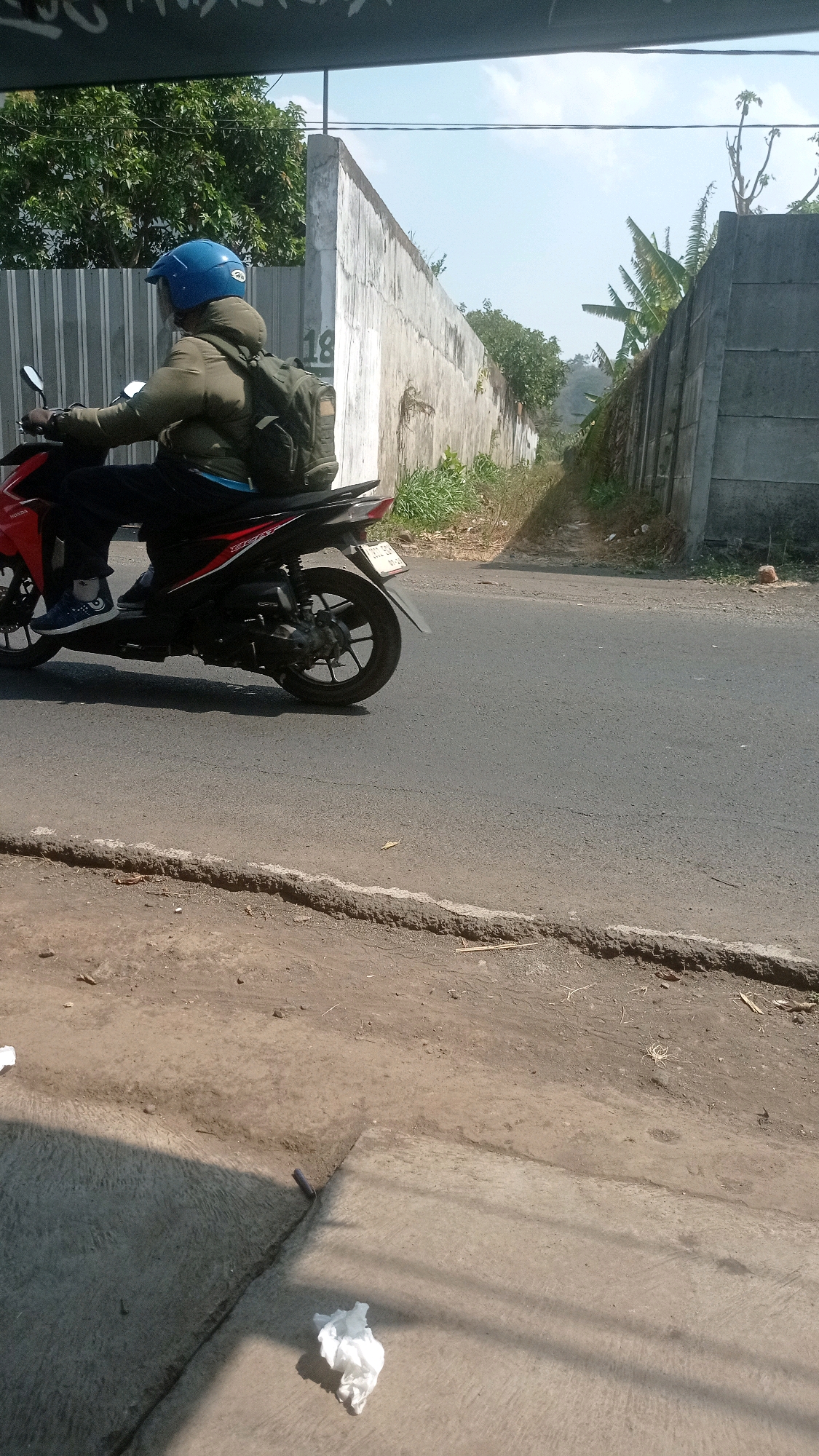 A person wearing a blue helmet and a khaki jacket rides a red and black motorcycle on a paved road in Bedali, Indonesia. The motorcycle has a license plate that reads "N 3434 BL". The rider is wearing dark pants and blue and white sneakers. A green backpack is strapped to the rider's back. The road is bordered by a dirt shoulder and a tall concrete wall. Lush green trees and vegetation are visible behind the wall. The sky is clear and blue. The scene appears to be during daylight hours.