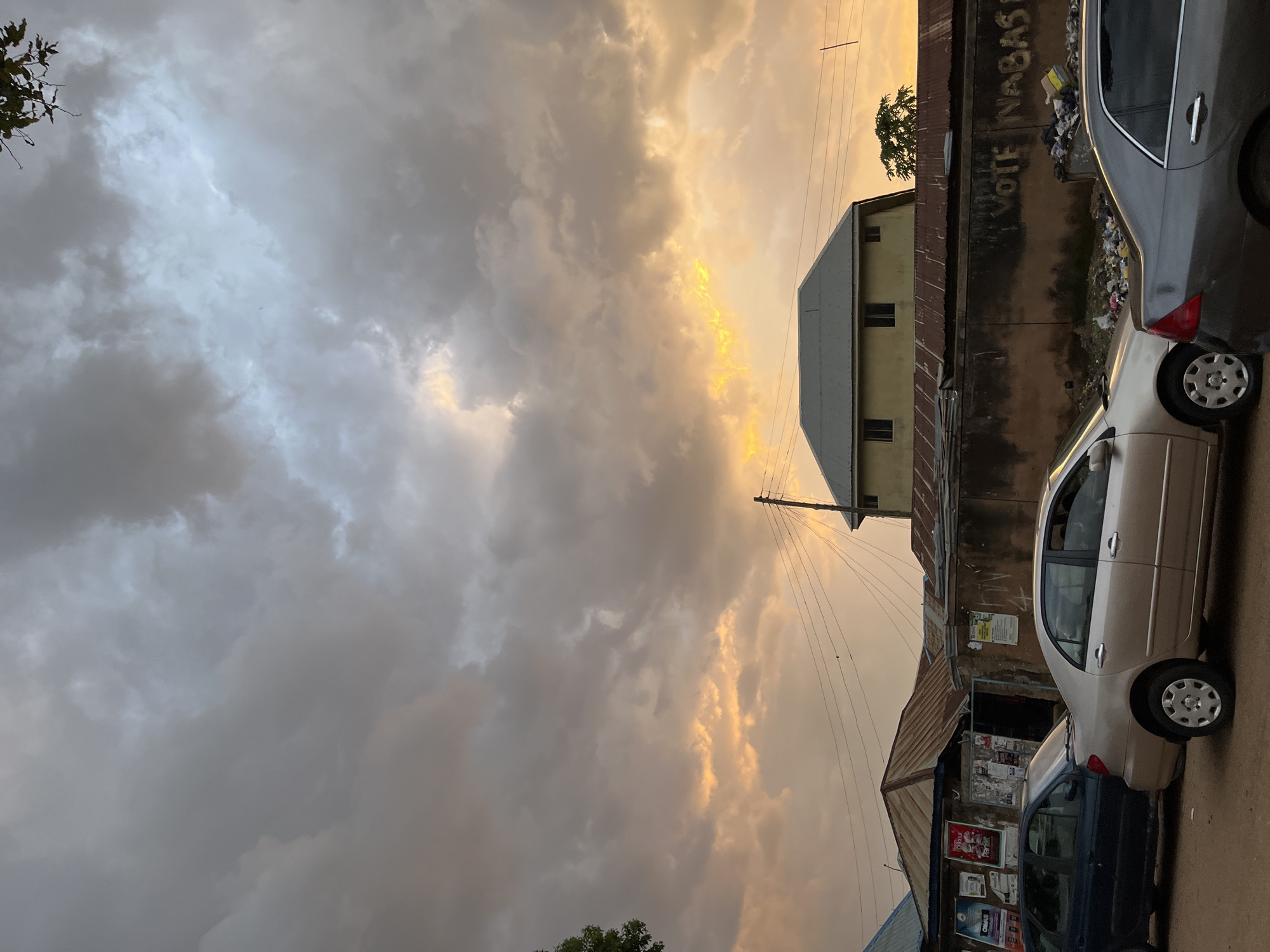 A street scene in Jos, Nigeria, is captured at dusk under a dramatic sky filled with heavy, grey clouds tinged with orange and yellow from the setting sun. In the foreground, three cars are parked along a dirt road: a dark blue sedan on the left, a light gold sedan in the center, and a dark gray sedan on the right. Behind the cars, a row of structures lines the street. A small shop with a corrugated metal roof and posters on its exterior is visible on the left. Further back, a larger building with a pale yellow facade and a grey metal roof stands prominently, with windows visible. A low, dark-colored wall runs across the midground, covered in graffiti that includes the words "VOTE NABAS." Piles of garbage and debris are scattered along the base of this wall. Electrical wires stretch across the scene, supported by a utility pole near the right edge of the frame. The overall impression is one of an urban setting experiencing a transition from day to night with an impending weather change.
