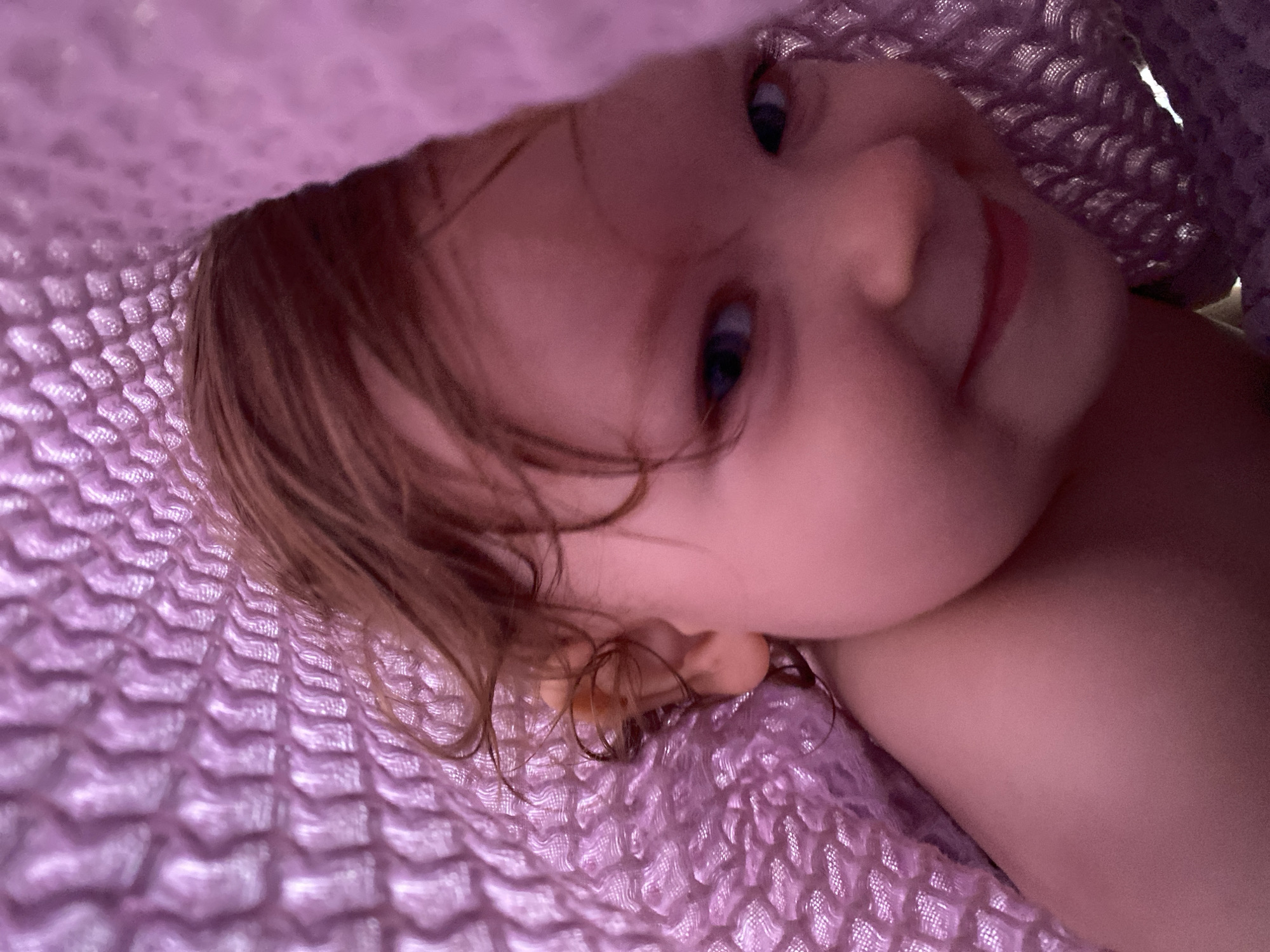 A close-up, canted shot depicts a young child partially covered with a textured, light purple blanket. The child has fair skin, light brown hair with soft curls near the ear, and light blue eyes. They are looking up and slightly to the right, exhibiting a faint smile. The blanket's texture is prominent and occupies a significant portion of the frame. Location information is unavailable.